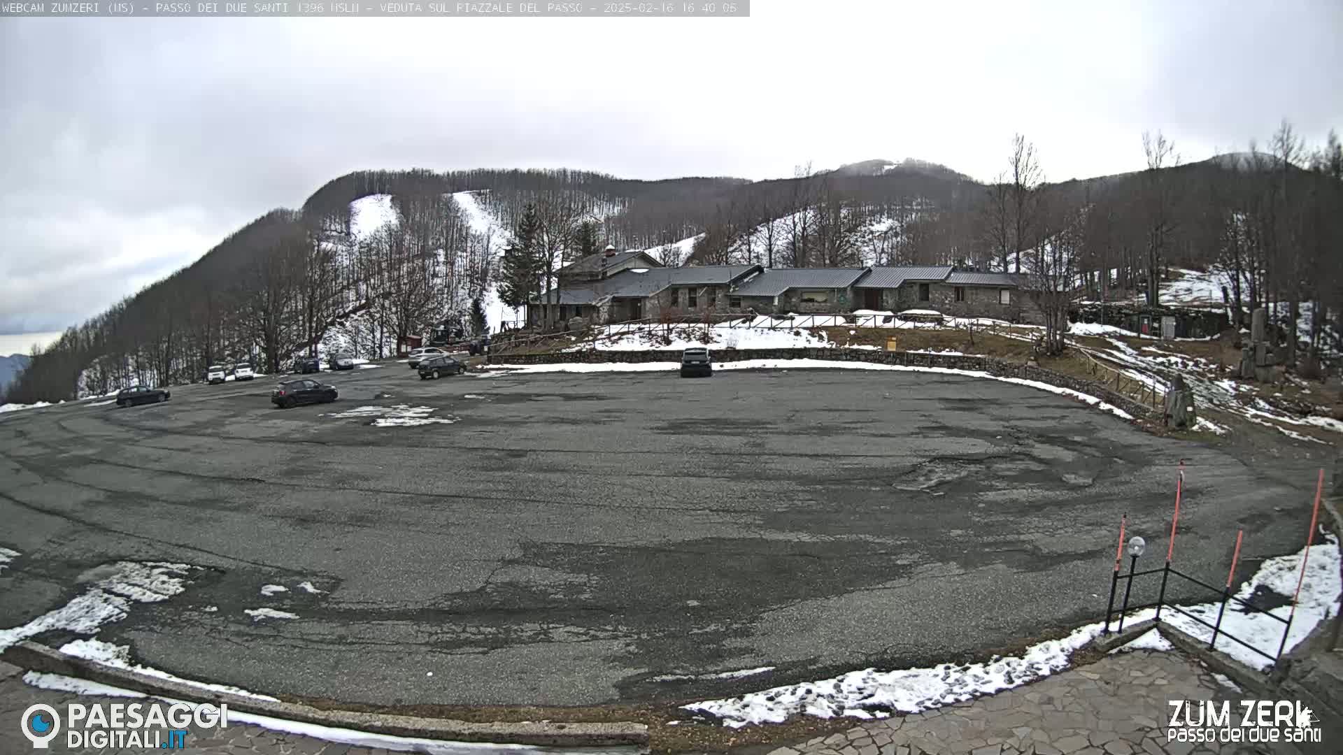A mostly empty asphalt parking lot is situated below a building and a snow-dusted mountainside under an overcast sky.