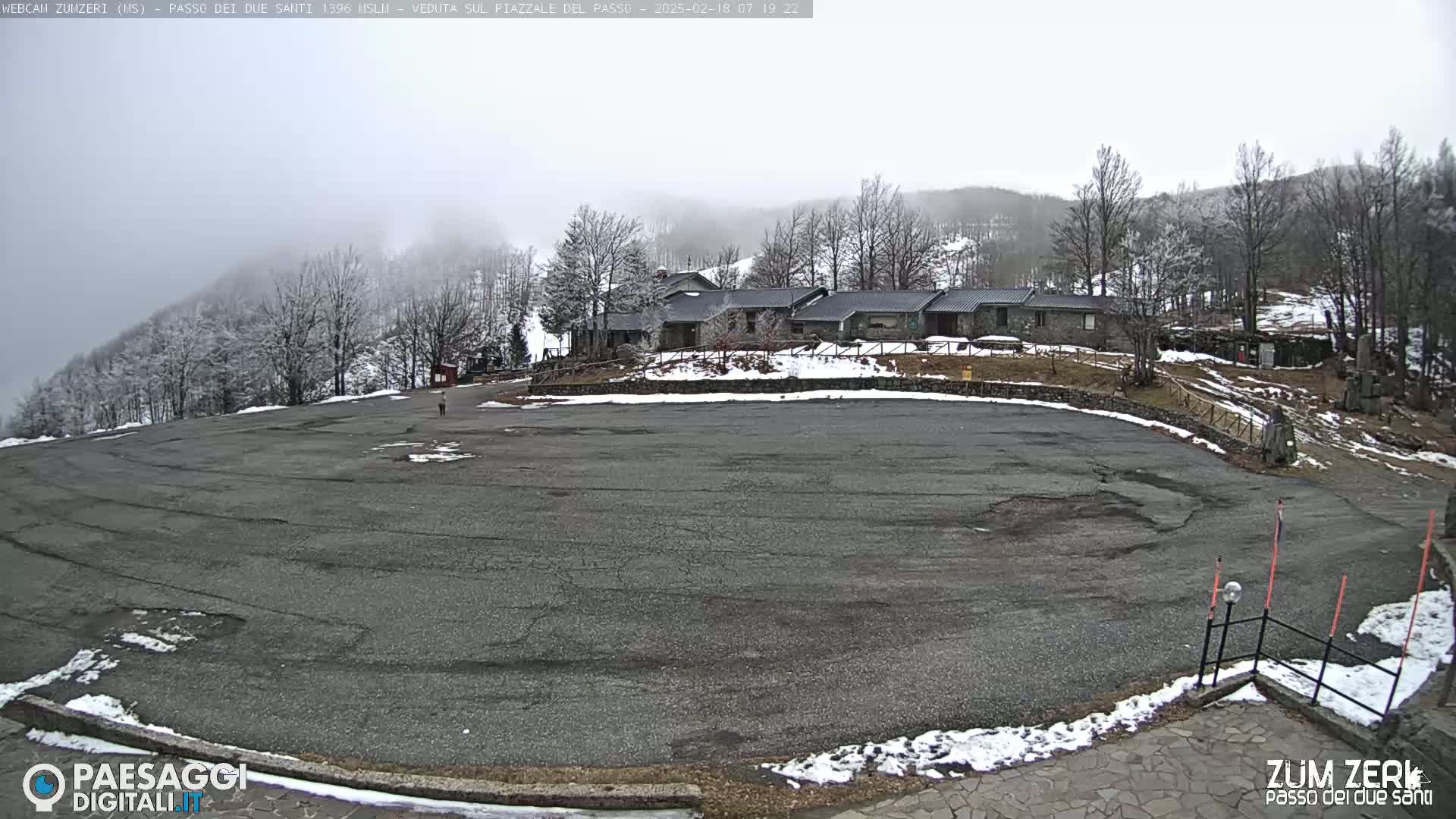 A large, empty asphalt parking lot sits in front of a stone building complex, partially covered in snow, with a hazy, overcast sky and snow-covered trees in the background.