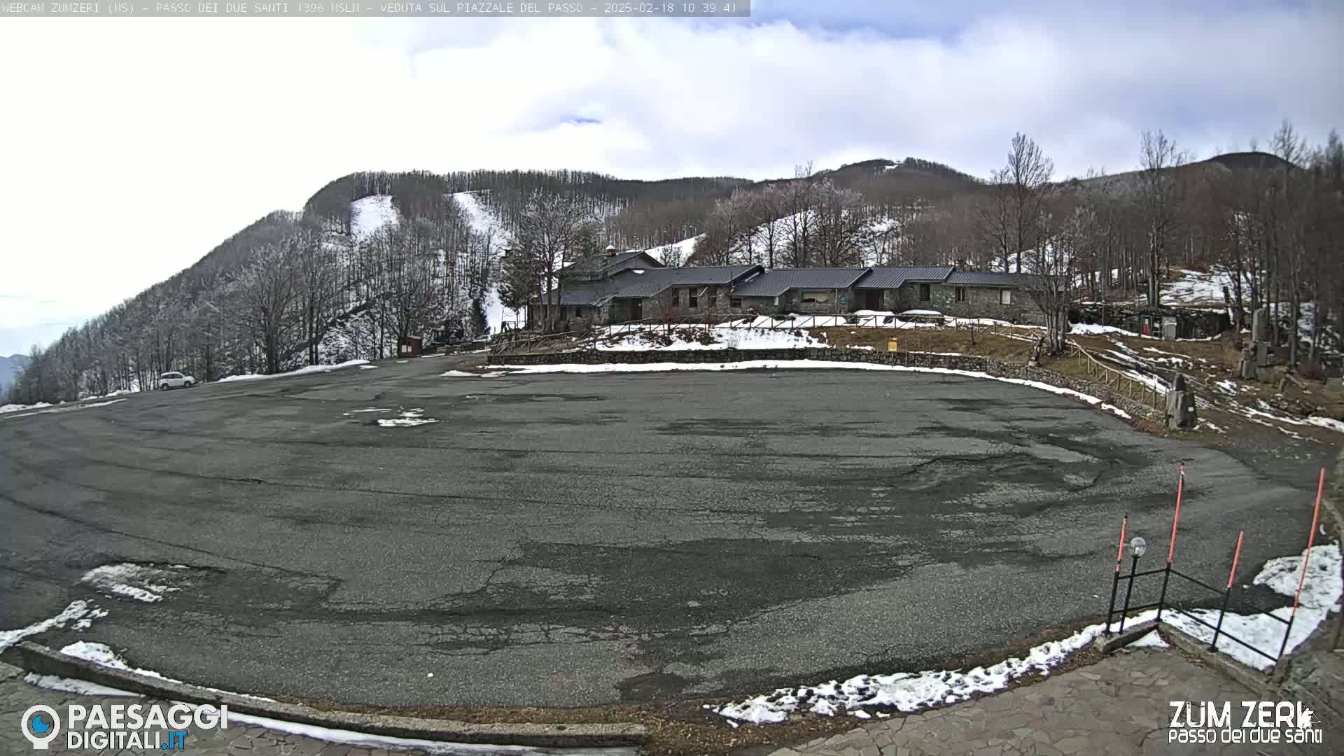 A mostly empty asphalt parking lot sits below a stone building complex nestled in a snow-dusted mountainside under a cloudy sky.