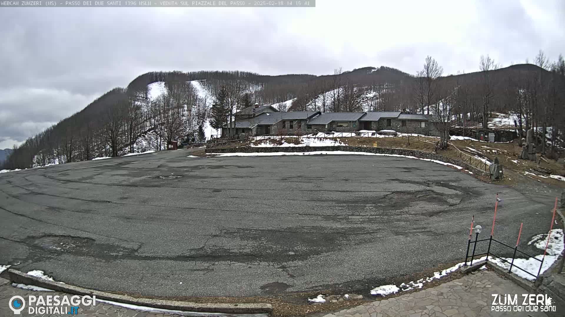 A large, empty asphalt parking lot sits in front of a stone building complex nestled amongst snow-dusted hills under a cloudy sky.