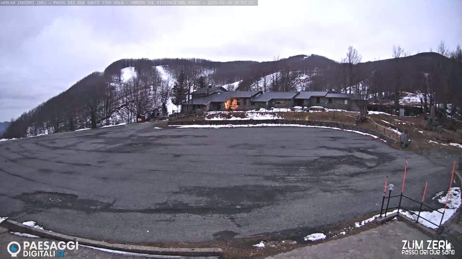 A large, empty asphalt parking lot sits in front of a stone building nestled in a snow-dusted mountainside under an overcast sky.
