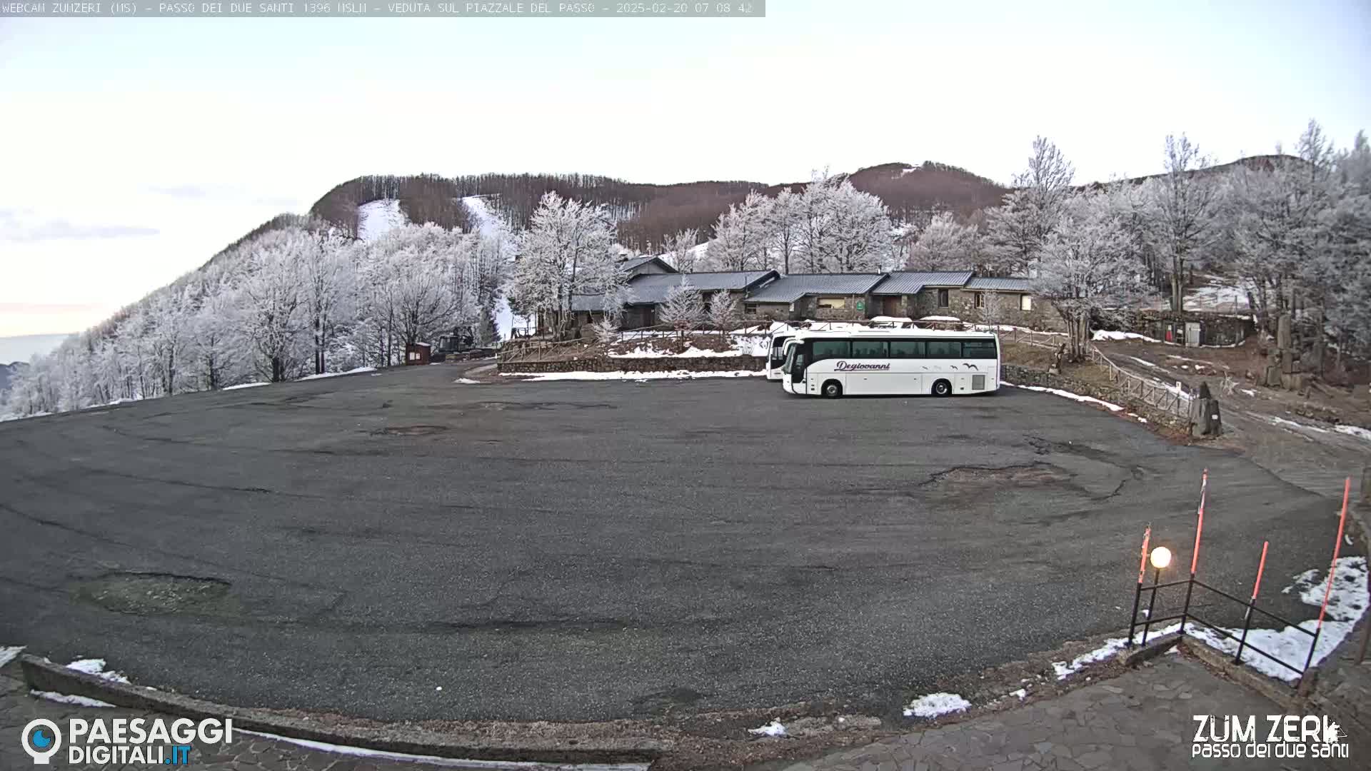 A large, empty asphalt parking lot sits in front of a building complex, with a few buses parked nearby, all nestled among frost-covered trees on a slightly snowy, overcast day.