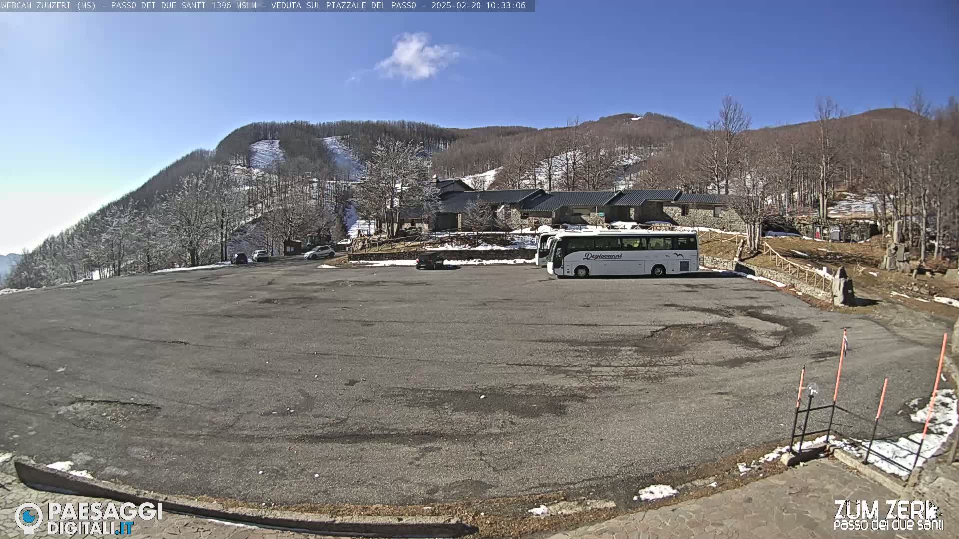 A mostly empty asphalt parking lot is situated in front of a building complex near a snow-dusted mountain under a clear blue sky.