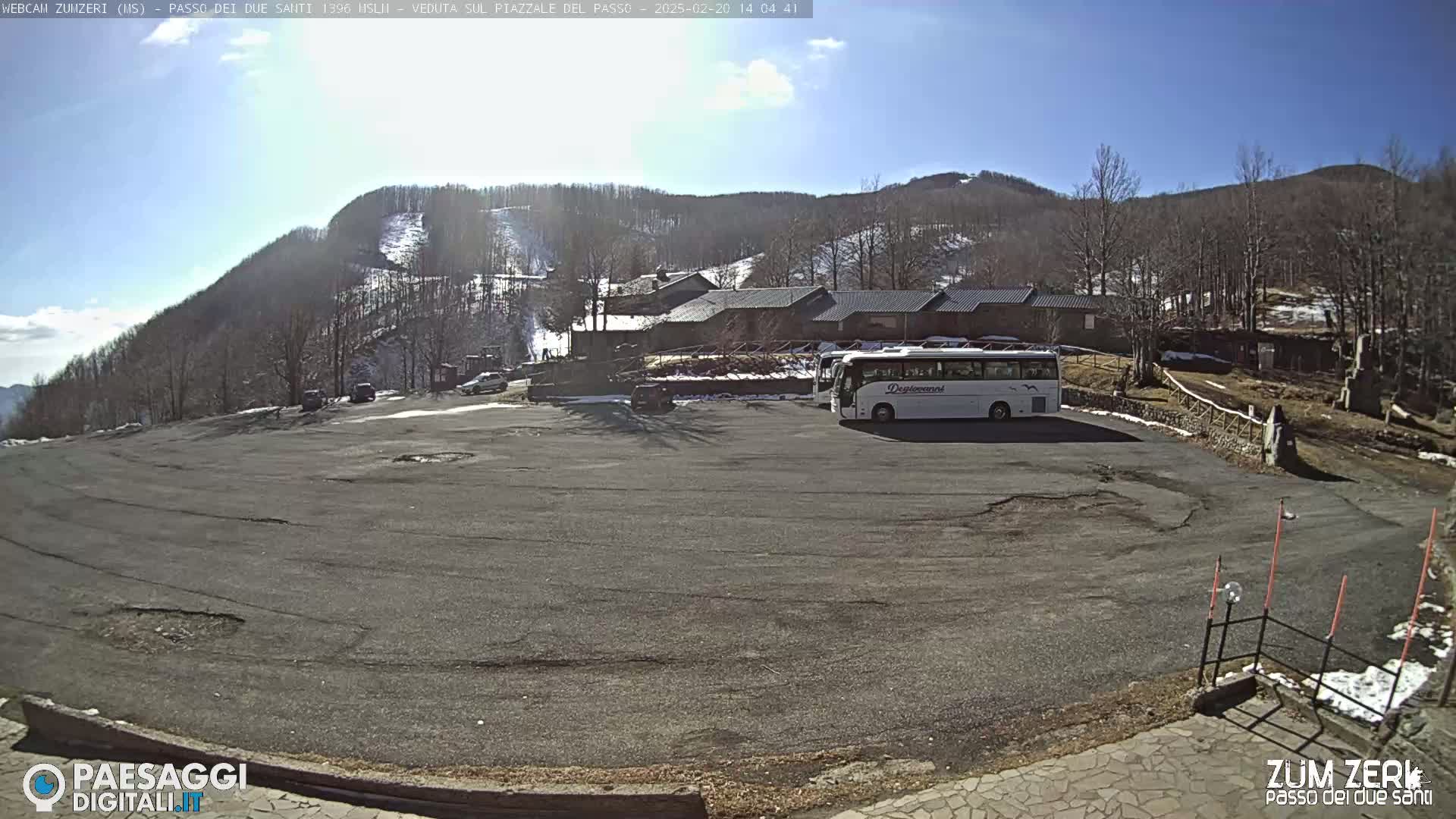 A mostly empty paved parking lot sits below a building and a snow-dusted mountainside under a clear, sunny sky.
