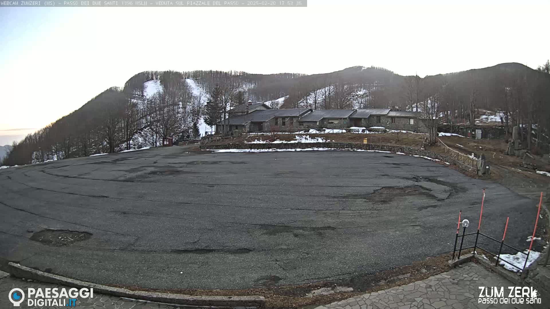 A large, empty asphalt parking lot sits in front of a stone building complex nestled among snow-dusted hills and trees under a mostly clear sky.