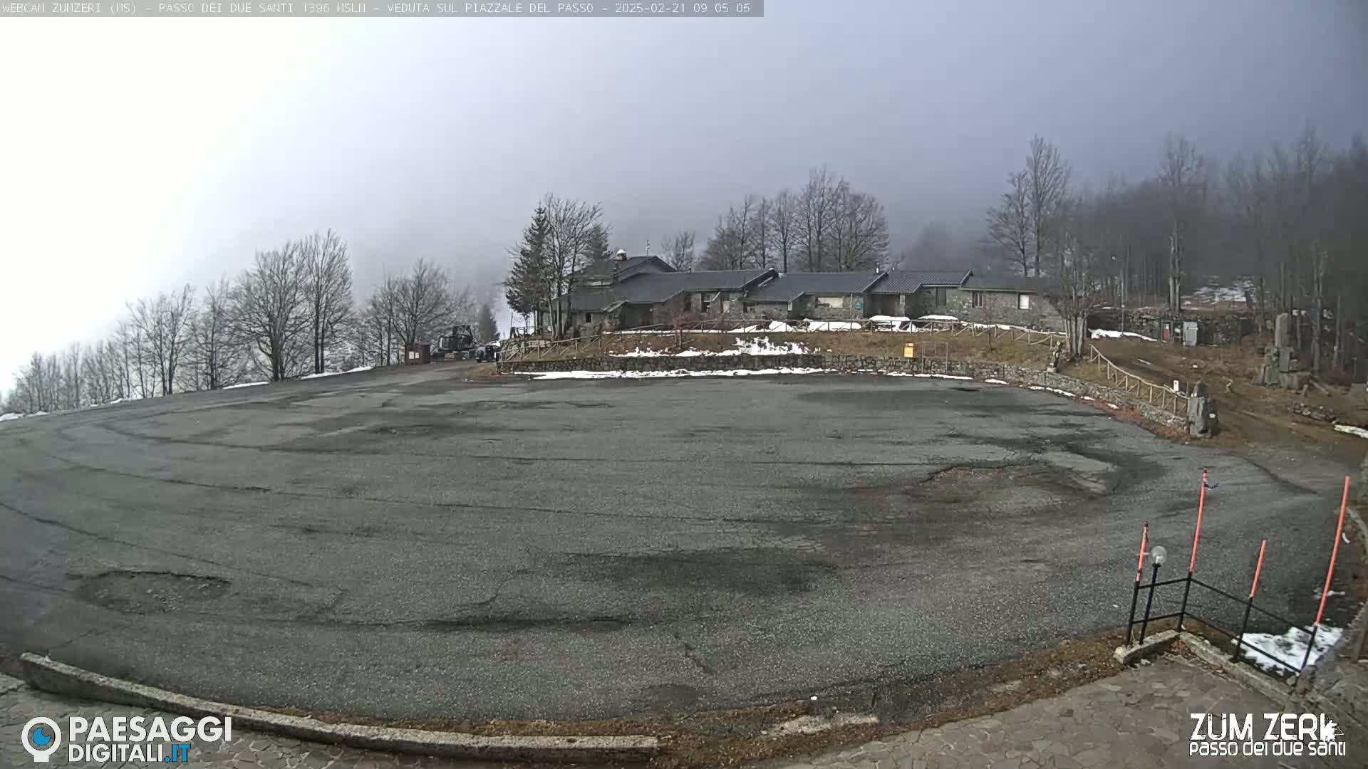 A large, empty asphalt parking lot is seen in front of a stone building complex, surrounded by leafless trees under an overcast sky.