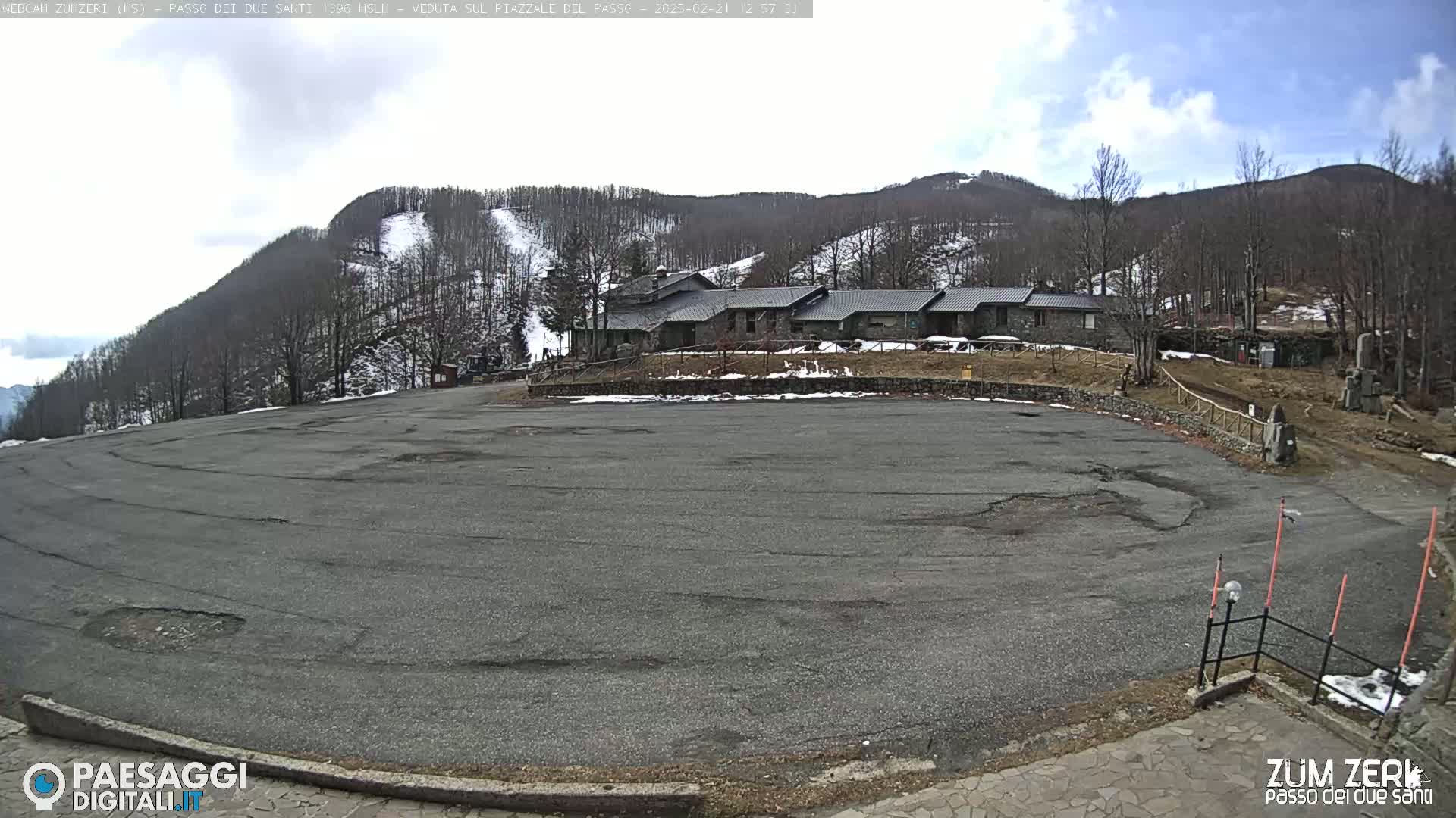 A large, empty asphalt parking lot sits in front of a stone building nestled in a snow-dusted mountainside under a partly cloudy sky.