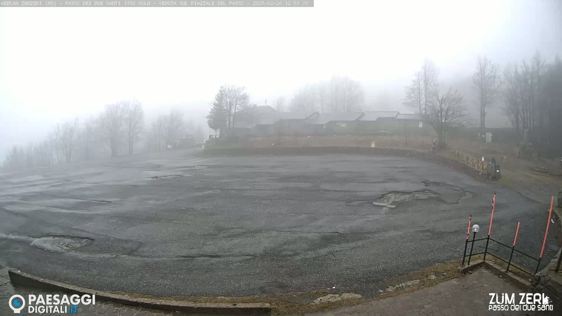 A large, empty asphalt lot with potholes is visible, partially obscured by a thick fog, and surrounded by bare trees and buildings in the distance.