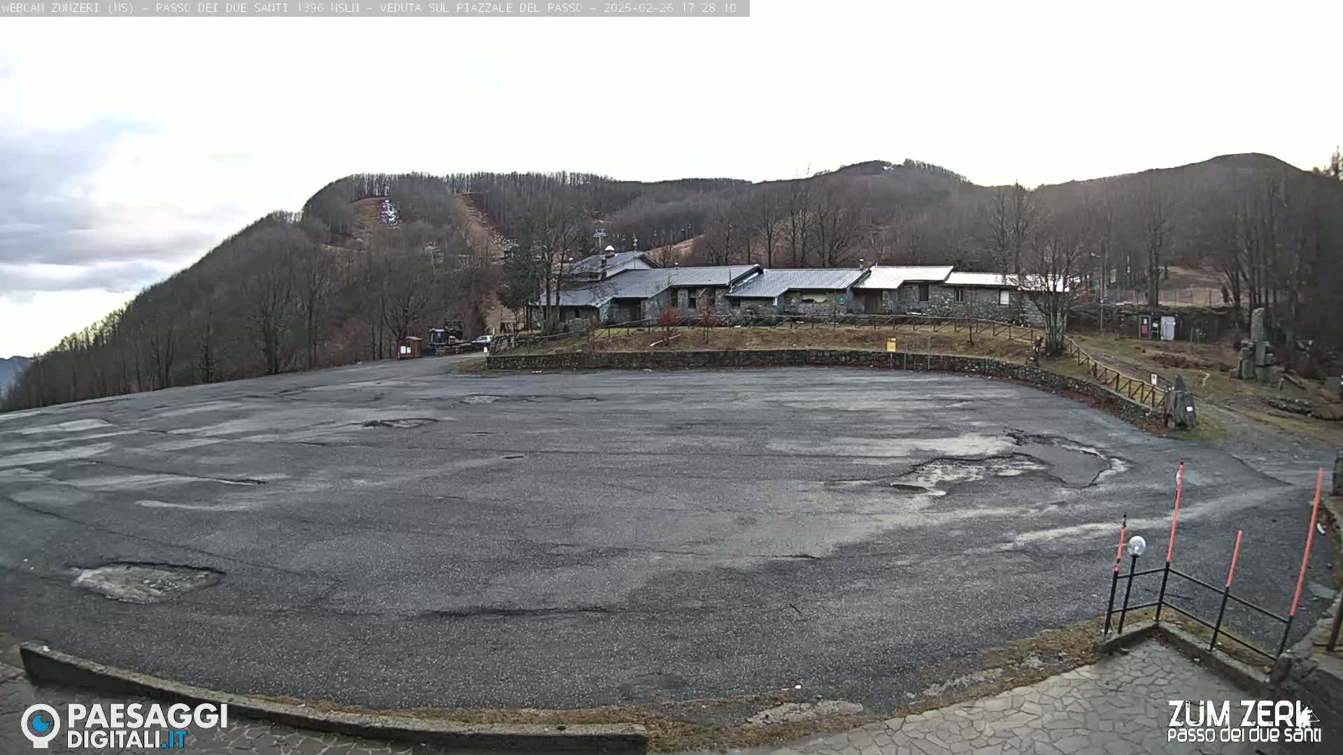 A large, cracked asphalt parking lot sits in front of a stone building complex nestled against a snow-dusted, treeline hill under an overcast sky.