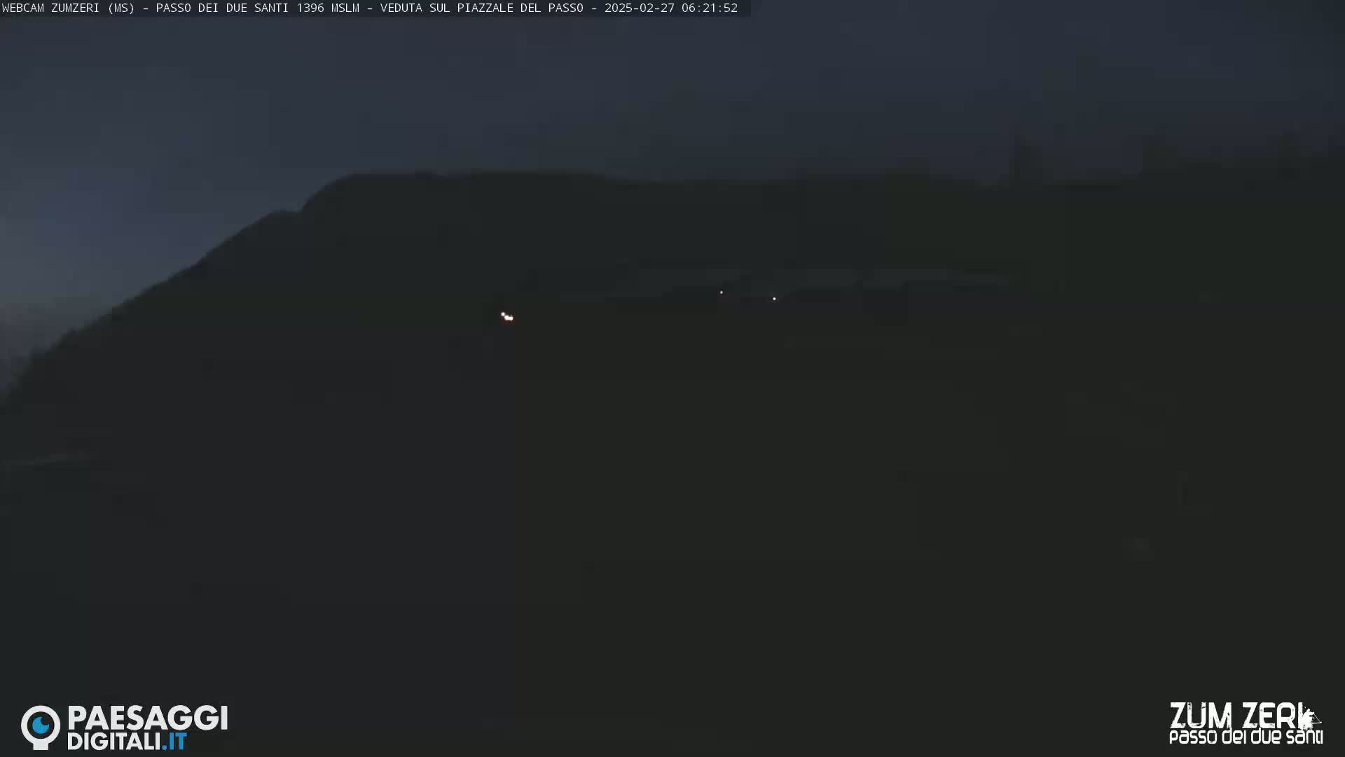 A dark, nighttime scene shows a hill in the distance with a few faint lights visible on a flat area in the foreground.