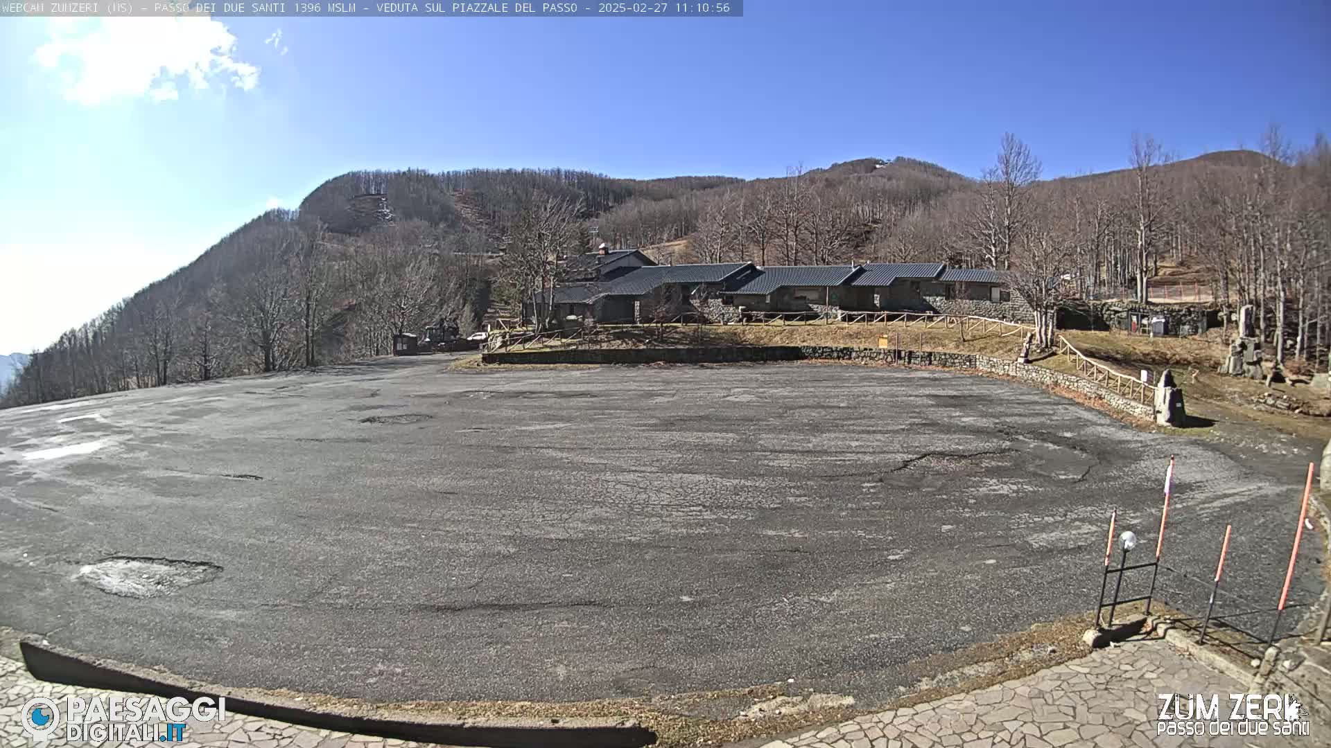 A large, empty asphalt parking lot is situated in front of a row of buildings, with a mountainside covered in leafless trees in the background under a clear, sunny sky.