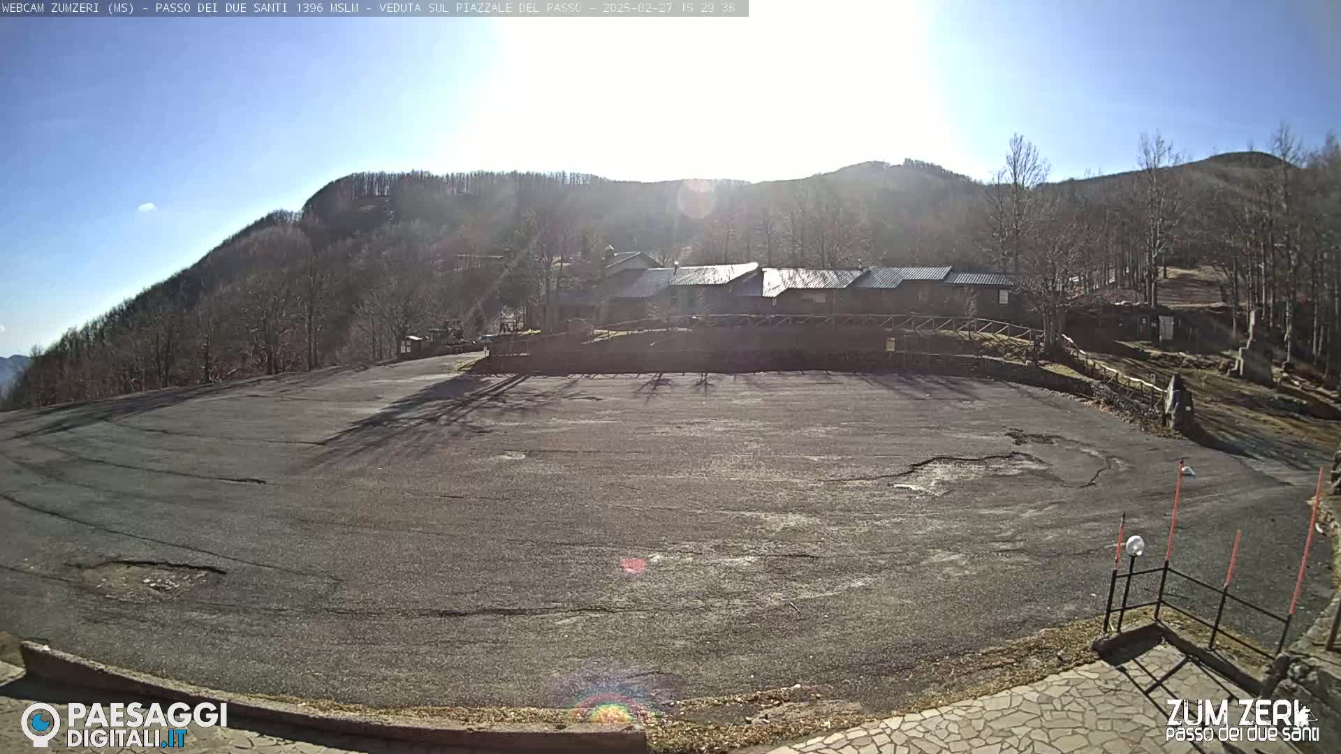 A mostly empty asphalt parking lot sits below a line of buildings against a backdrop of a sunny, snowless mountain covered in bare trees.