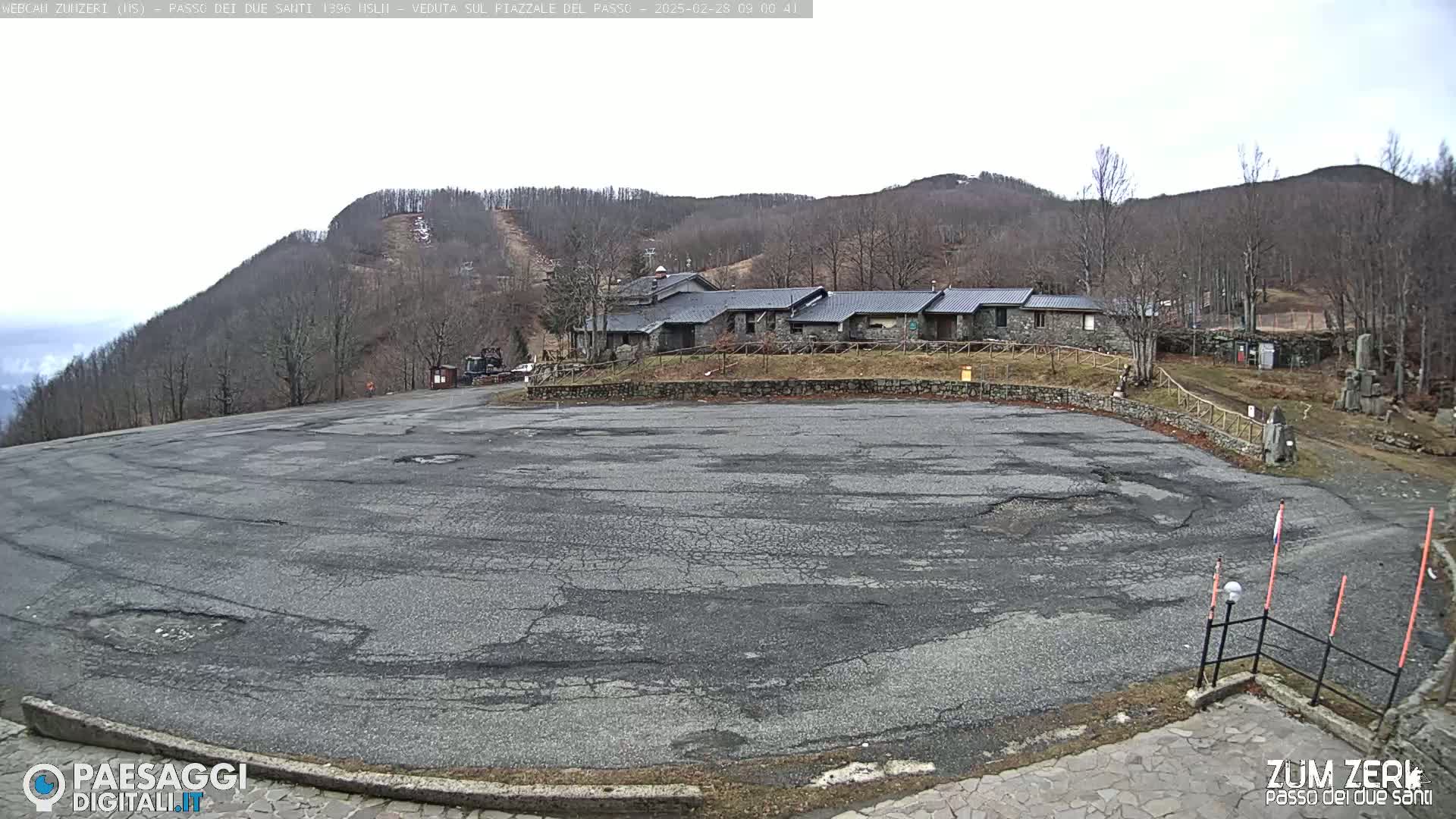A large, cracked asphalt parking lot sits in front of a stone building complex nestled against a hillside covered in leafless trees under an overcast sky.