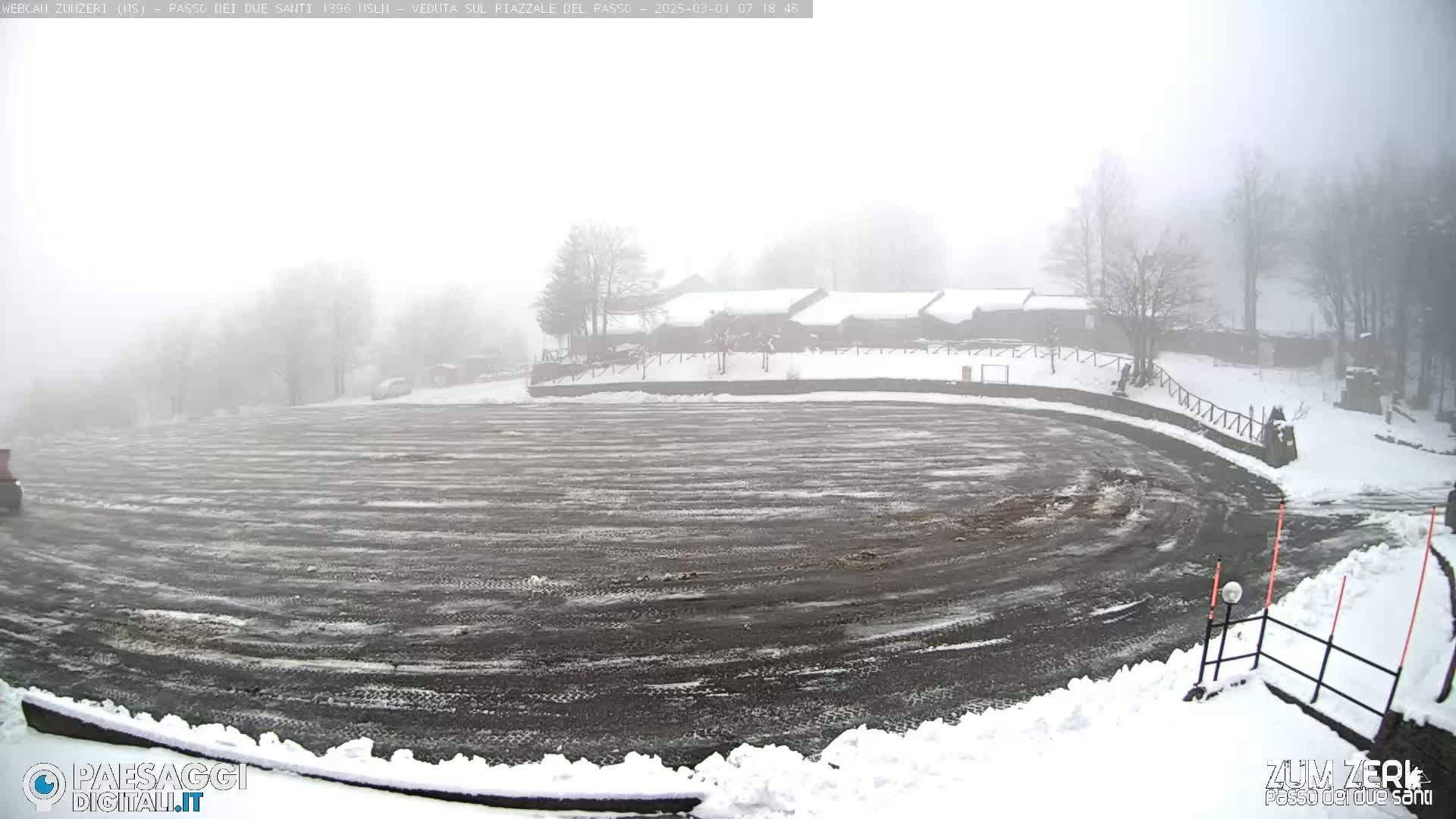 A snow-covered, empty parking lot is visible in foggy conditions, with a few buildings and trees in the background.
