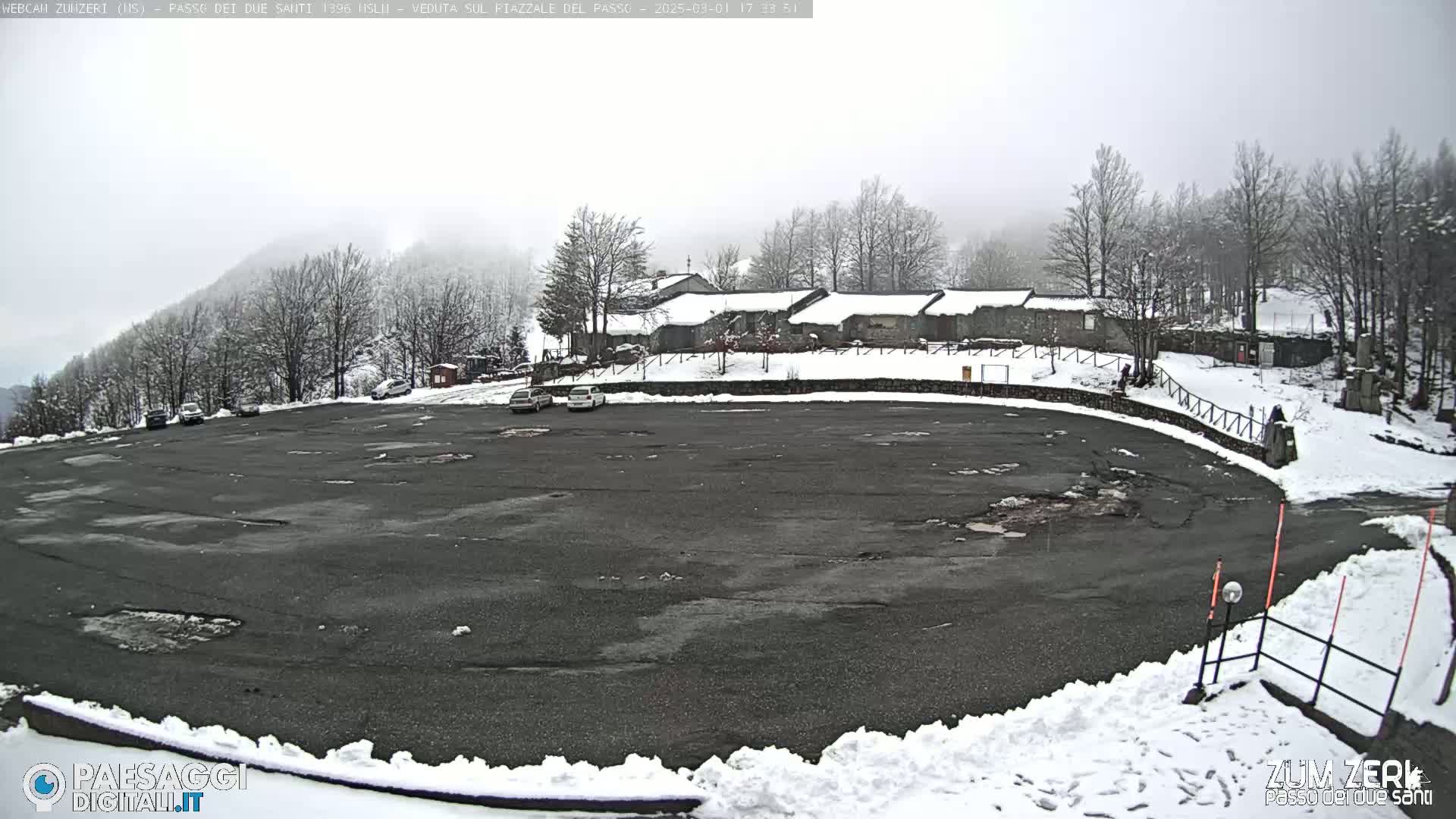 A mostly empty asphalt parking lot is surrounded by snow, with a few cars parked and a building in the background under an overcast sky.