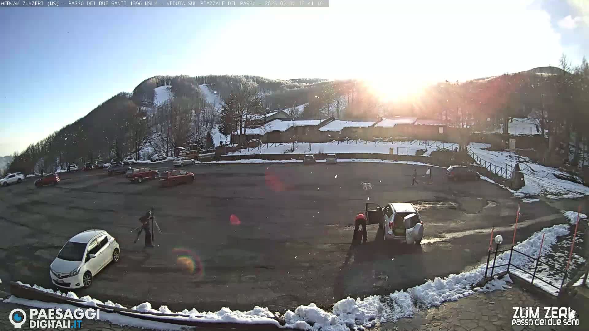 A snow-dusted parking lot with several cars and people, situated below a sunlit mountain with a ski slope visible on its side.
