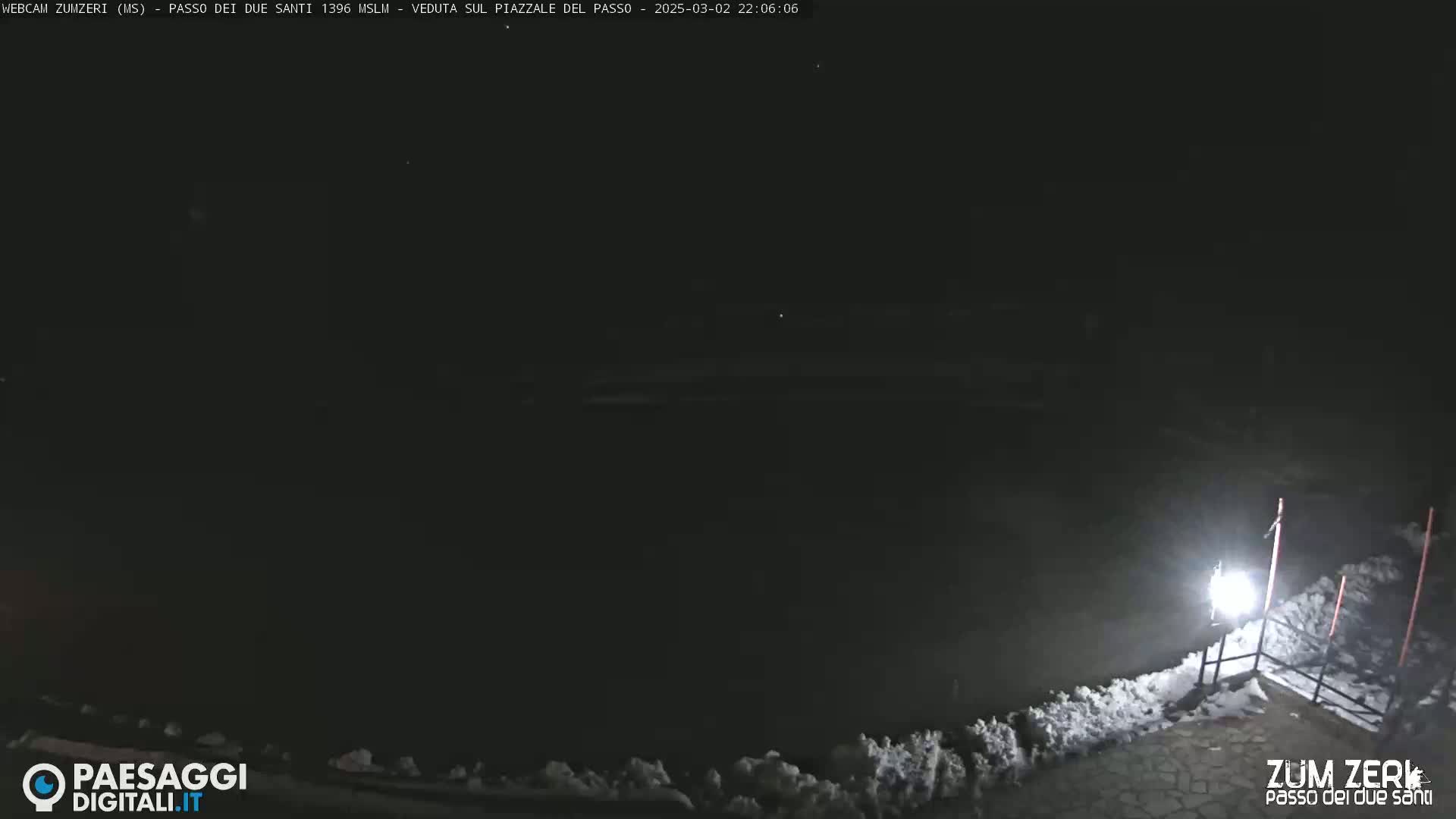 A nighttime, snow-covered scene is illuminated by a single bright light, showing a dark expanse beyond a low fence.