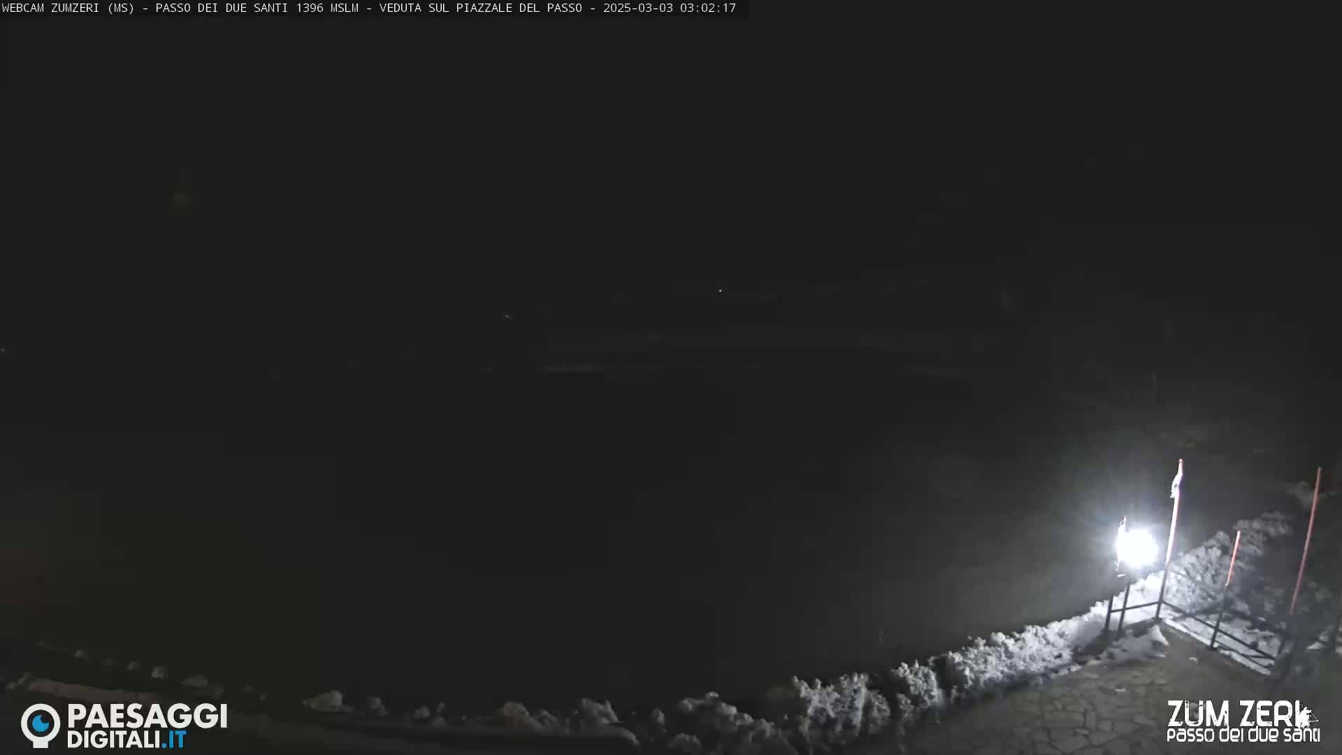 A dark nighttime scene shows a snow-covered area with a single lit lamppost near a railing.