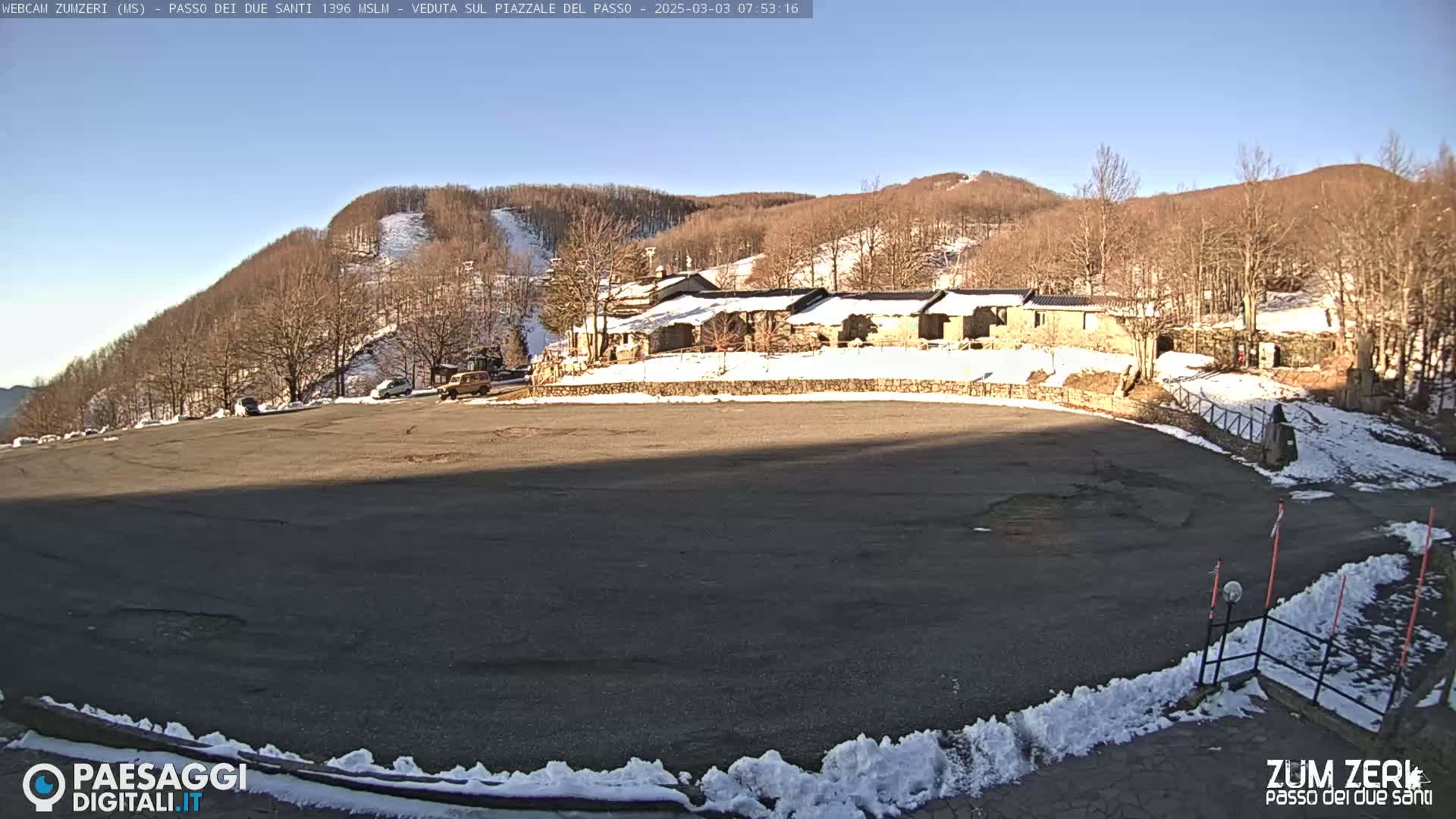 A large, mostly empty asphalt parking lot is situated in front of a cluster of stone buildings, with snow-covered hills and leafless trees forming a backdrop under a clear, sunny sky.