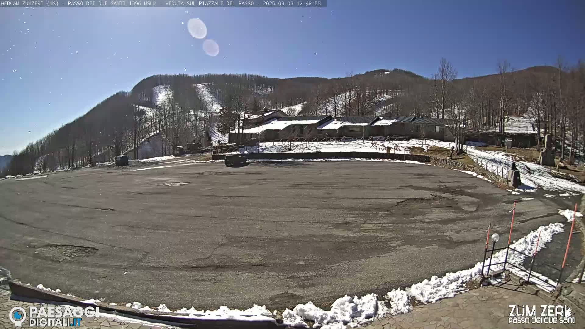 A large, mostly empty asphalt parking lot is visible with a few patches of snow, overlooking a snow-dusted mountainside under a clear blue sky.