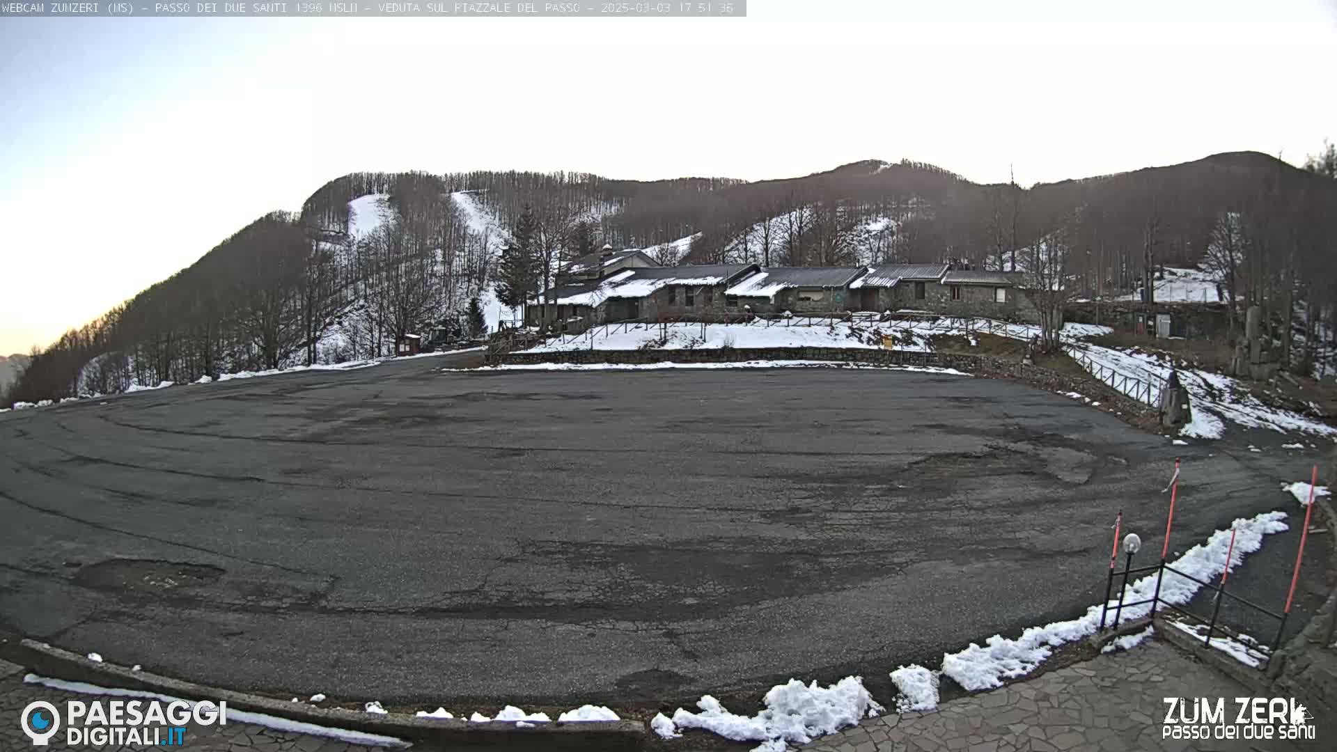 A large, empty asphalt parking lot sits before a cluster of buildings nestled at the base of a snow-dusted mountain under a clear sky.
