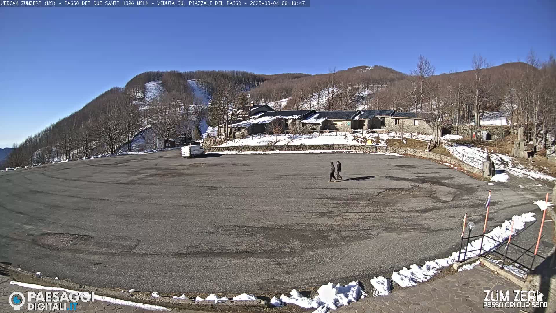 A large, mostly empty asphalt parking lot is situated in front of a stone building complex, with snow on the ground and in the surrounding, sparsely wooded hills under a clear blue sky.