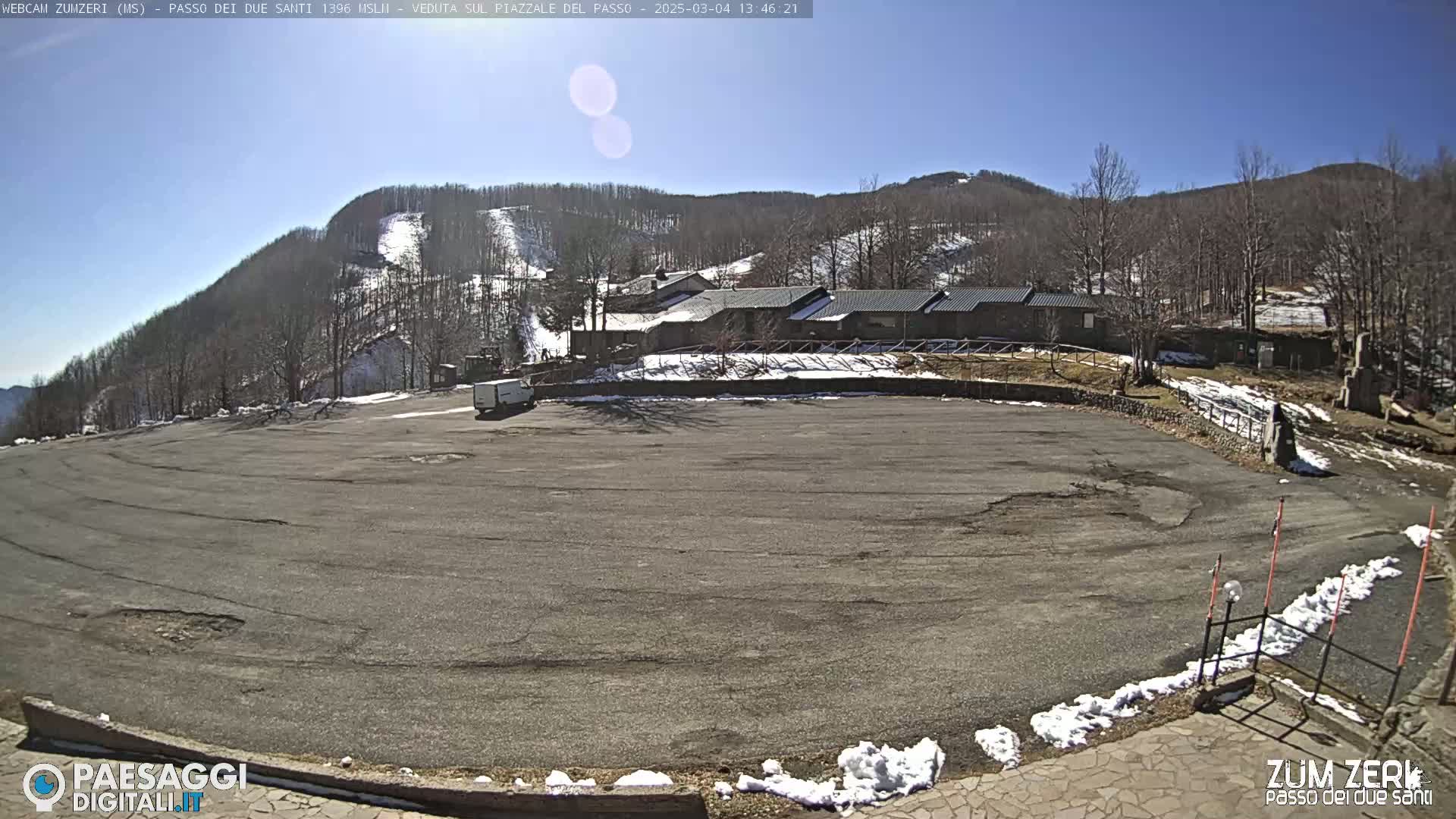A large, empty asphalt parking lot sits below a building near a snow-dusted mountainside under a clear, sunny sky.