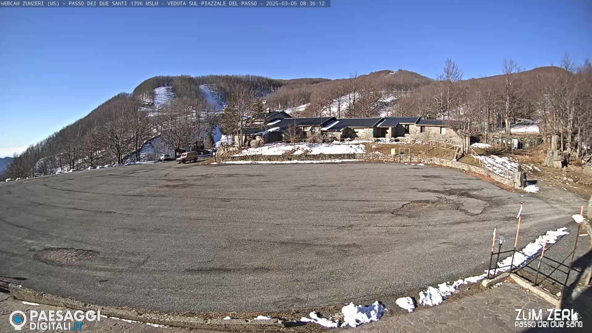 A large, mostly empty asphalt parking lot sits below a stone building complex nestled in a snow-dusted mountainside under a clear blue sky.