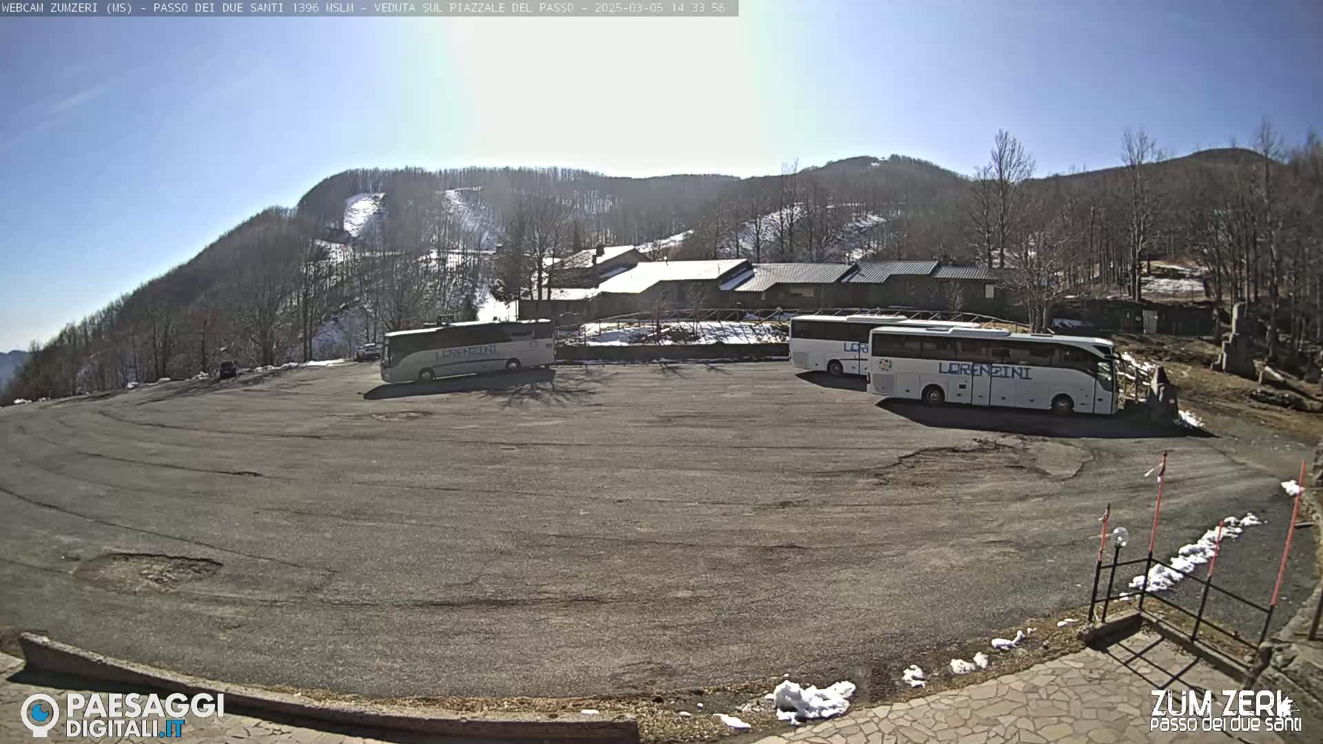 A sunny day reveals several buses parked in a lot overlooking a mountain with sparse snow and leafless trees.