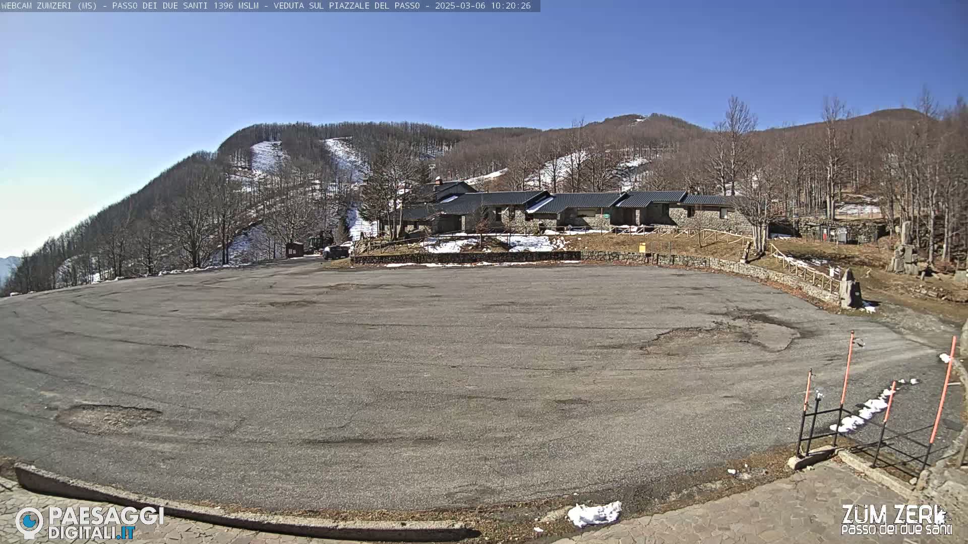 A large, empty asphalt parking lot is situated before a cluster of low stone buildings, with a snow-dusted hillside rising behind them under a clear blue sky.