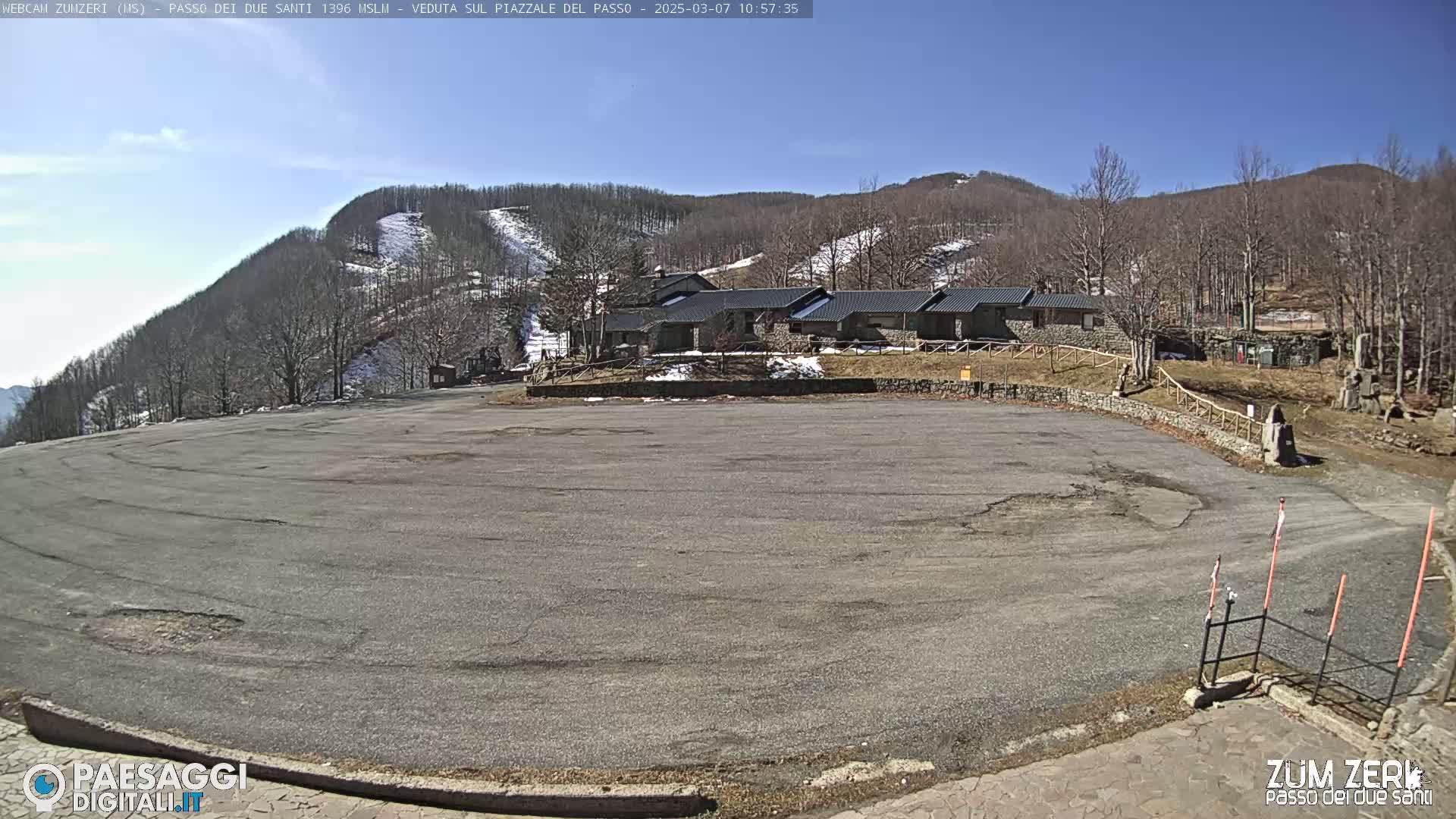 A large, empty asphalt parking lot is situated in front of a cluster of stone buildings, with snow-dusted hills and a mostly clear blue sky in the background.