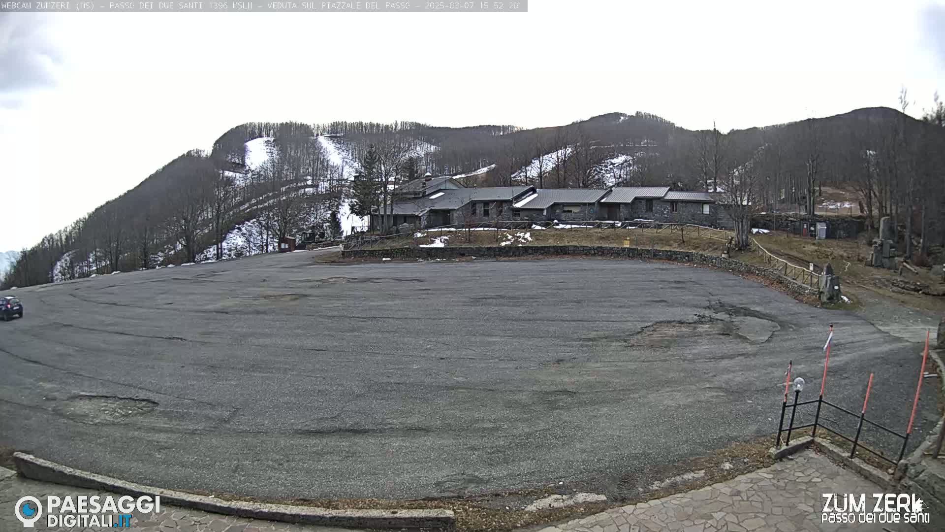 A large, empty asphalt parking lot sits before a row of stone buildings, which are nestled at the base of a snow-dusted mountain under a cloudy sky.