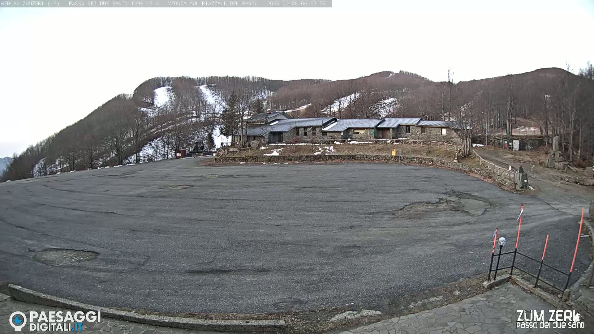 A large, empty asphalt parking lot sits before a stone building complex nestled at the base of a snow-dusted mountain under a cloudy sky.