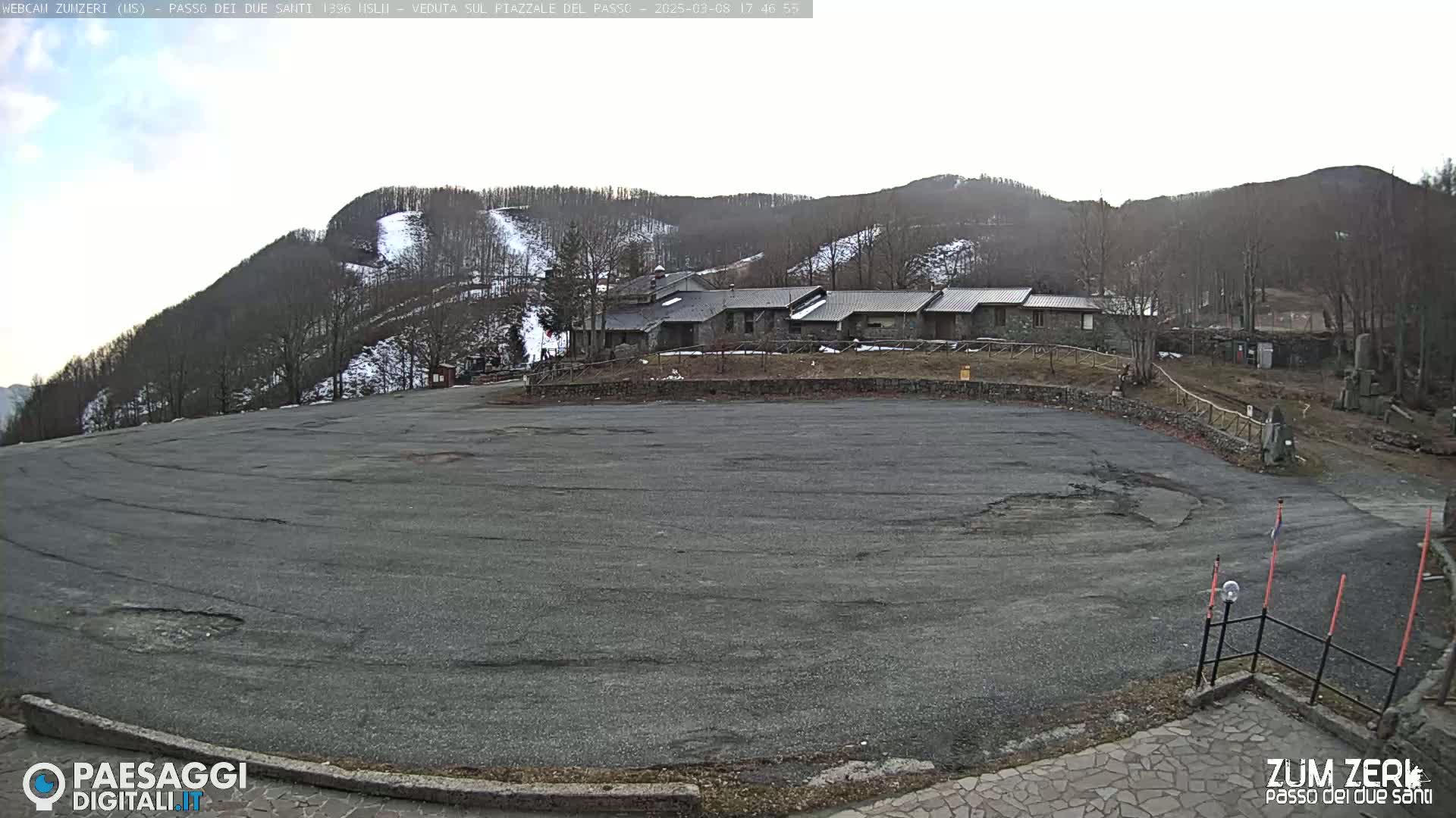 A large, empty asphalt parking lot sits below a stone building, overlooking a snow-dusted hillside under a mostly clear sky.