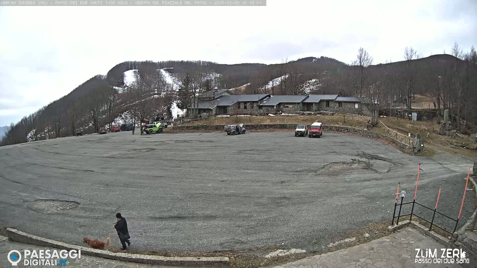 A mostly empty, large asphalt parking lot sits below a building complex nestled in a snow-dusted mountainside under an overcast sky.