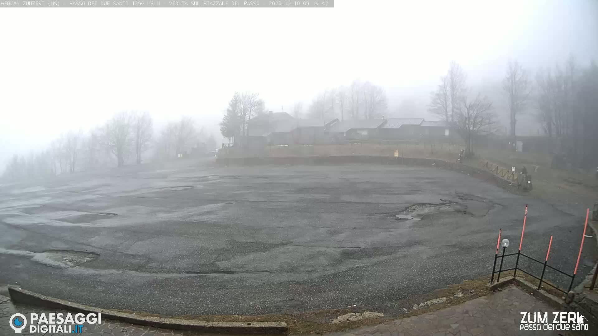 A foggy, overcast day reveals a large, empty asphalt parking lot with some buildings and trees in the background.