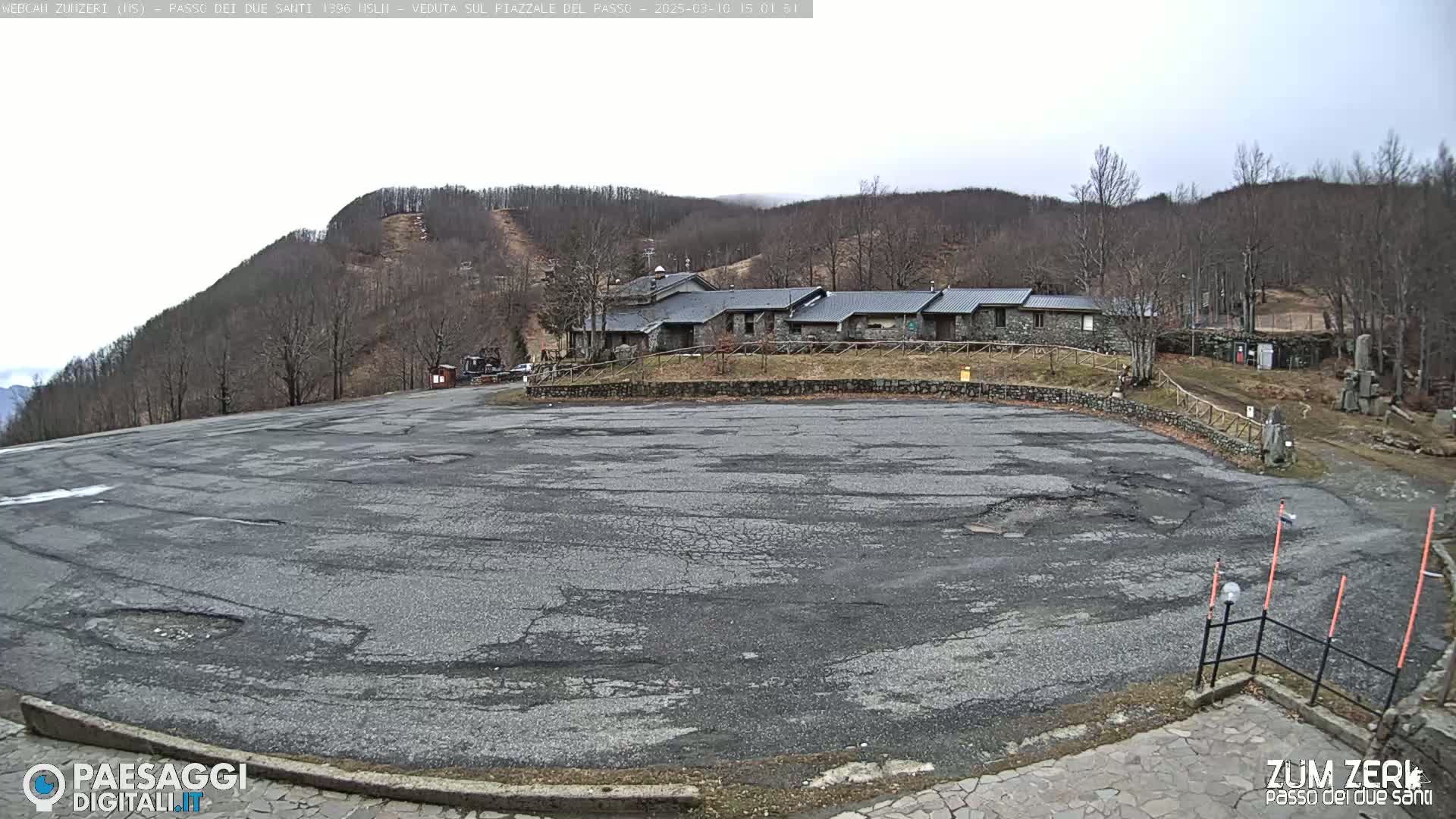 A large, cracked asphalt parking lot sits before a stone building complex nestled among bare, winter trees on a hillside under an overcast sky.