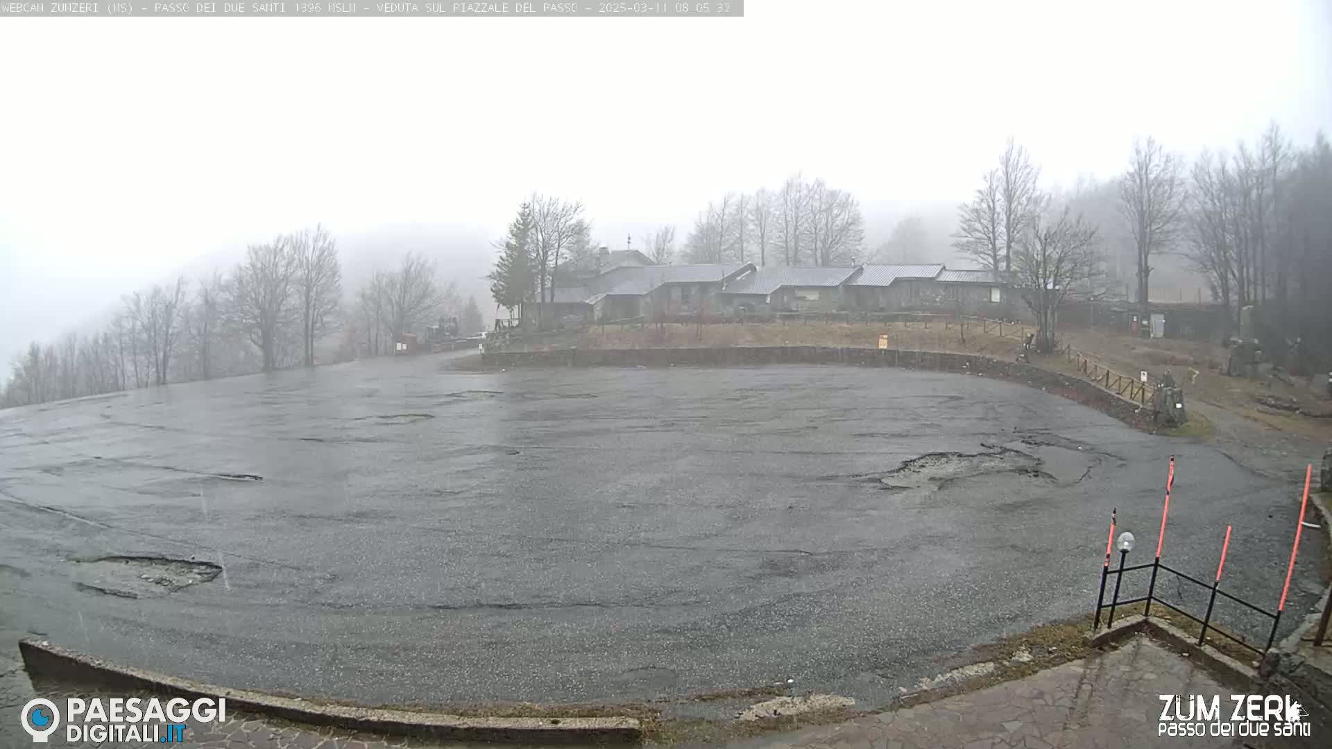 A large, empty, rain-soaked asphalt parking lot sits in front of a cluster of low buildings under a foggy, overcast sky.