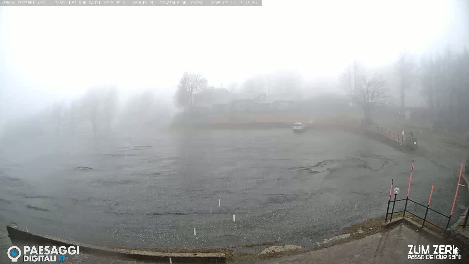 A foggy, rainy scene shows a mostly empty parking lot with a few trees and buildings barely visible in the distance.