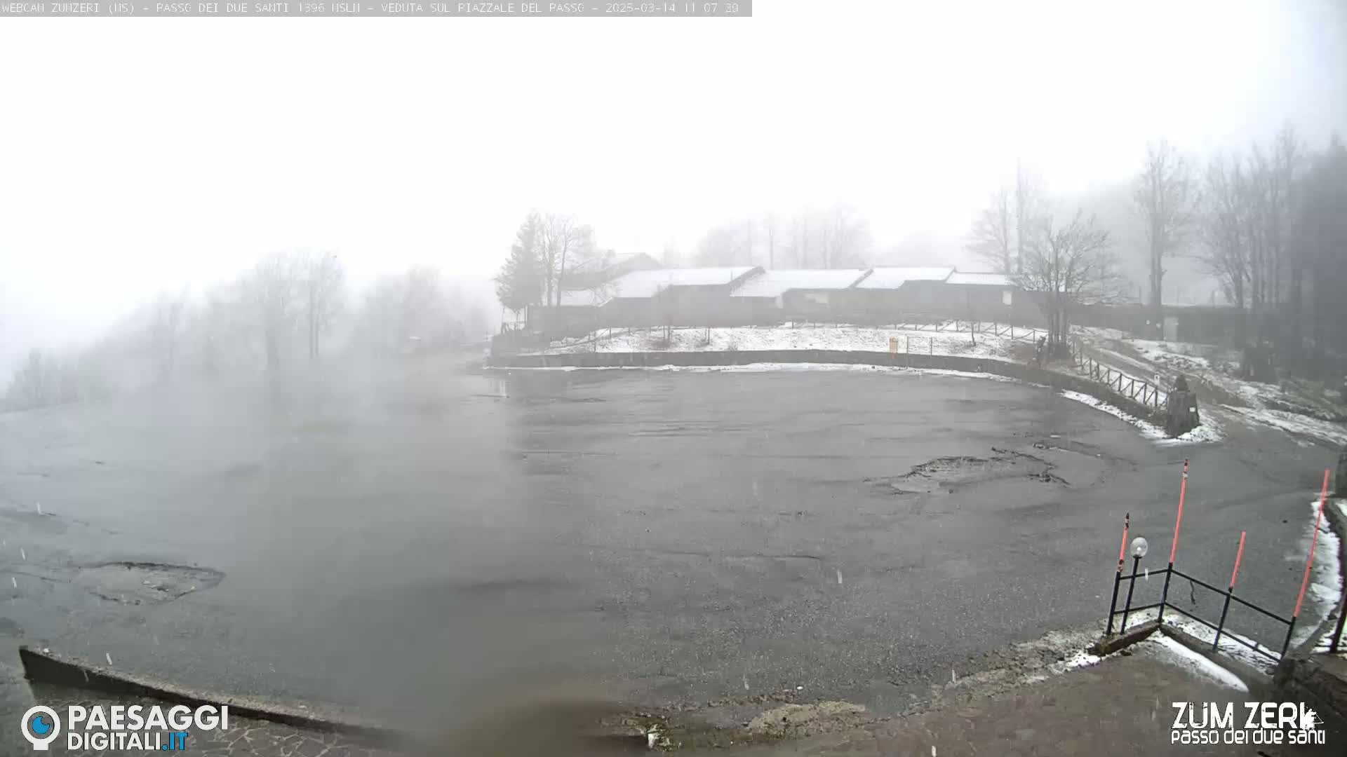 A snow-covered parking lot and building are visible through a heavy fog or mist with light snowfall.