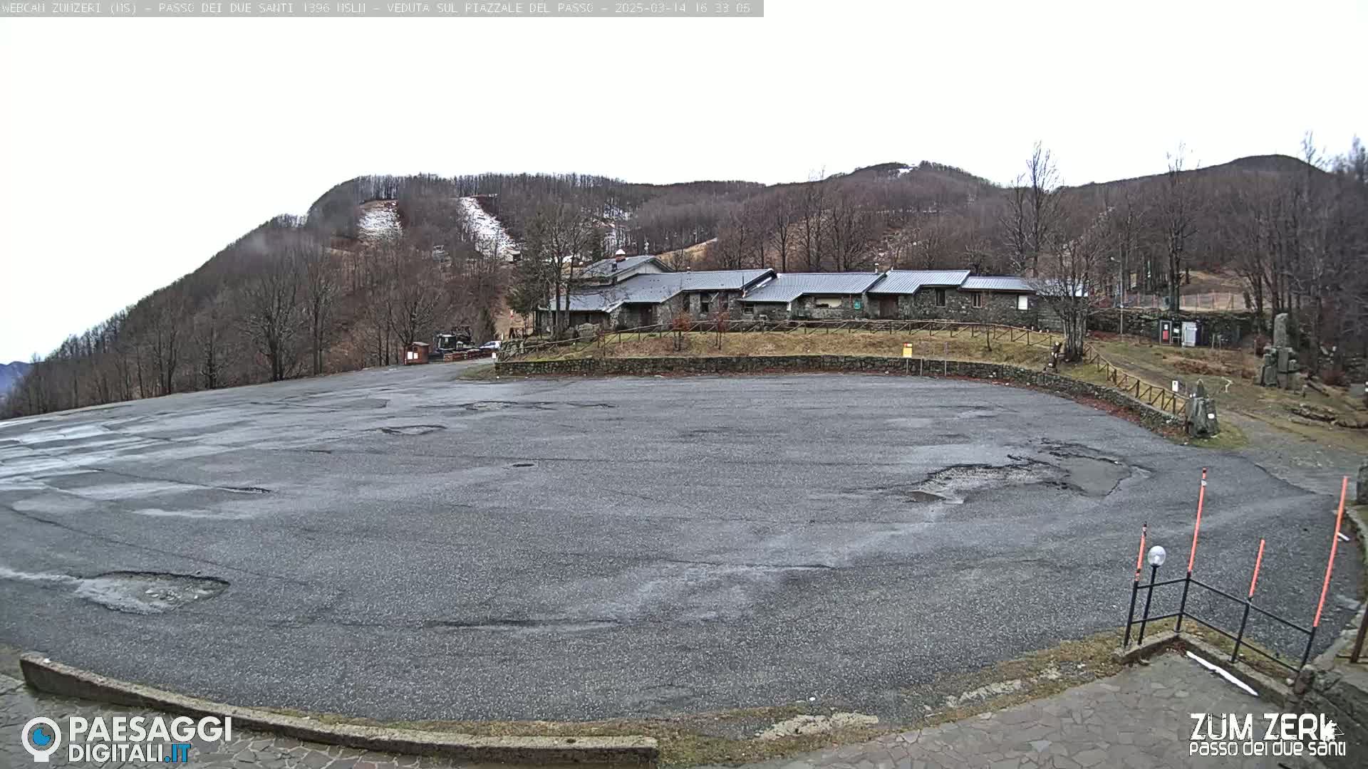 A large, empty asphalt parking lot with potholes is situated in front of a stone building complex, nestled amongst bare, winter trees on a hillside under an overcast sky.