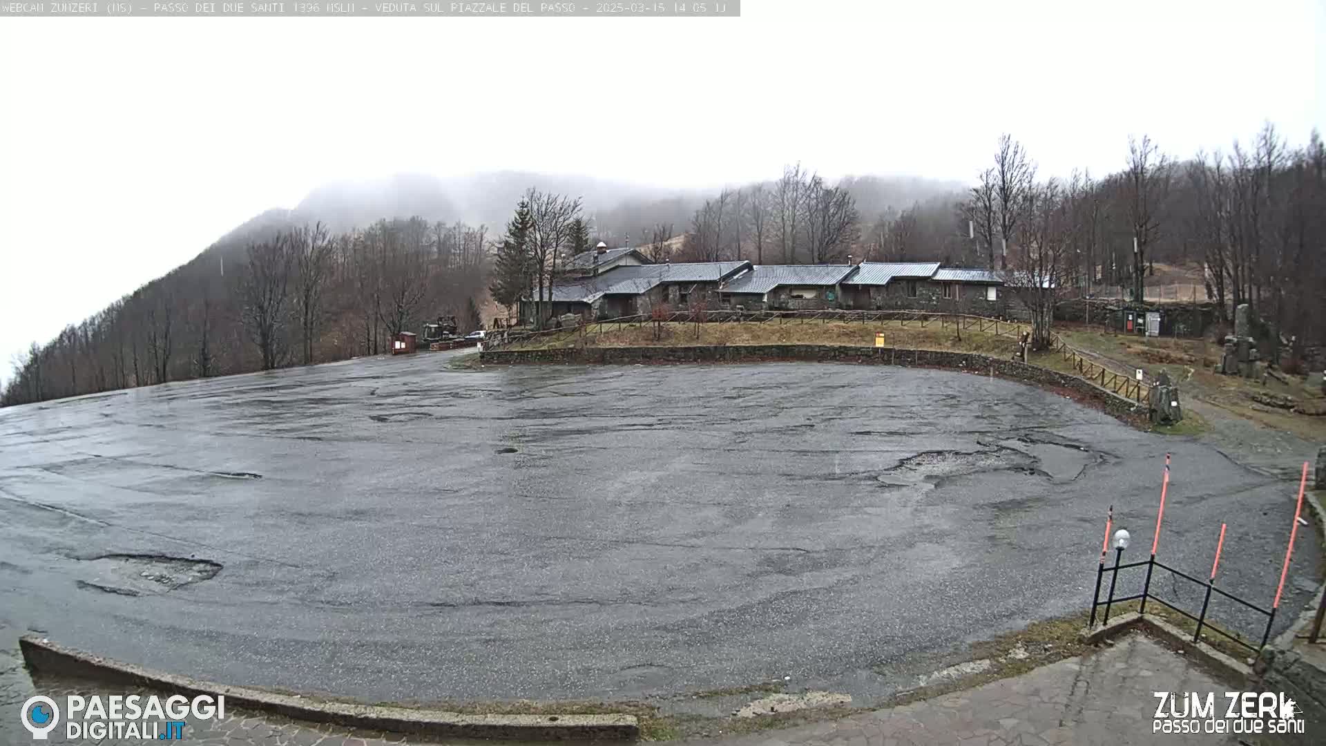 A wet, empty asphalt parking lot sits in front of a stone building complex, with misty, leafless trees and hills in the background.