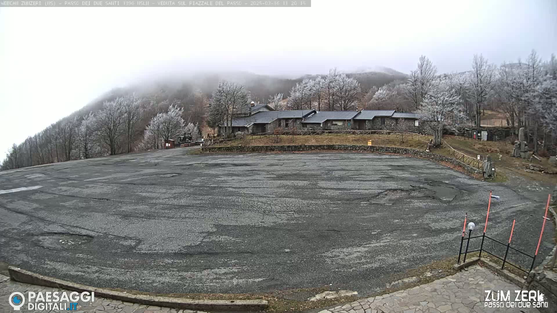 A large, empty asphalt parking lot sits in front of a stone building complex nestled amongst frost-covered trees on a foggy mountainside.
