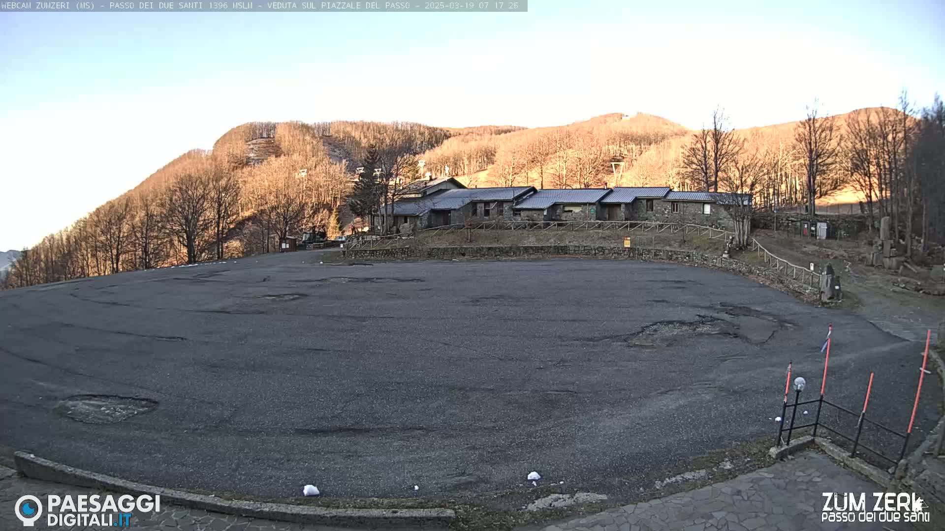 A large, empty asphalt parking lot sits before a row of stone buildings, which are nestled at the base of a hill covered in bare, brown trees under a clear, bright sky.