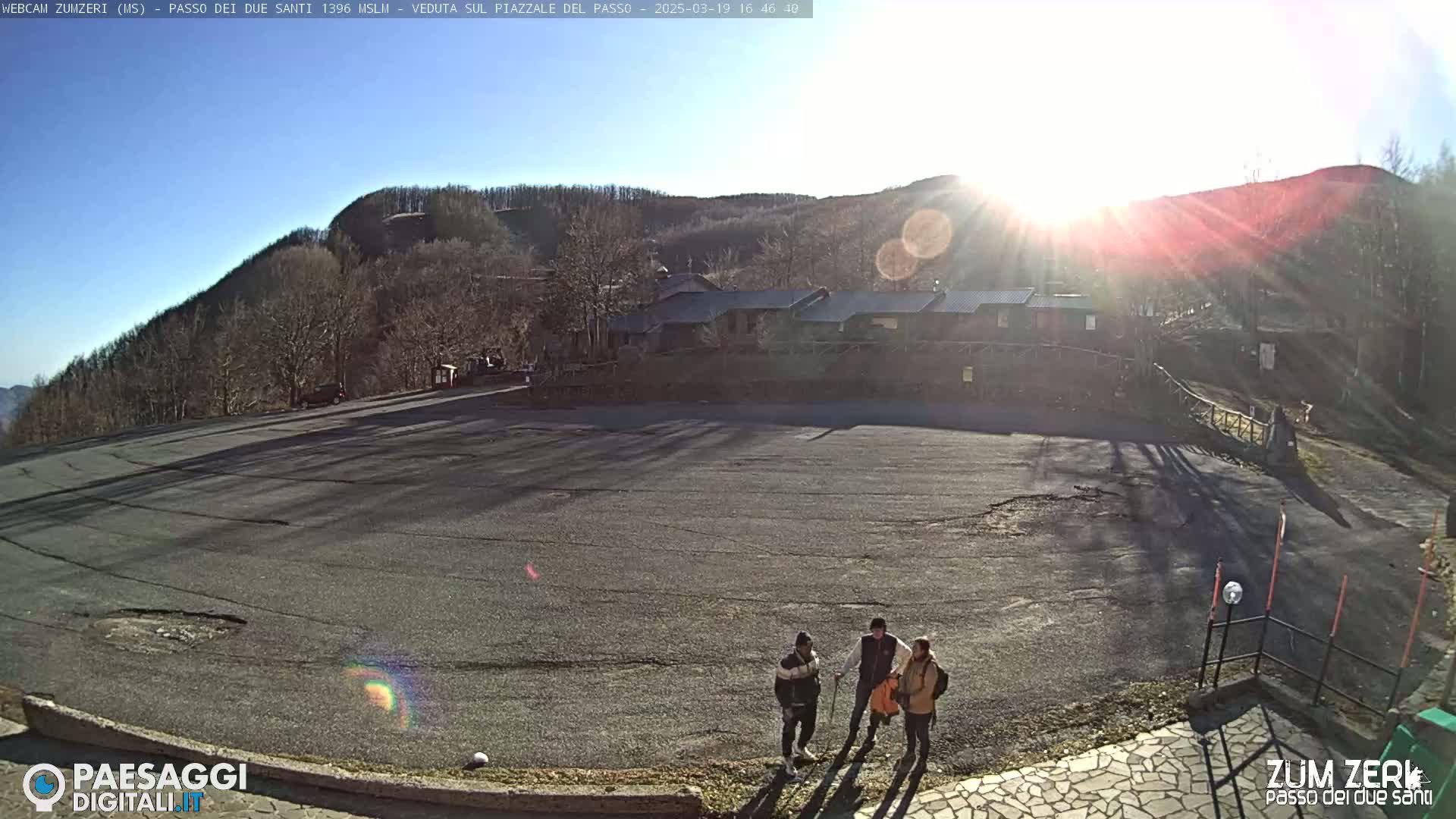 A sunny, outdoor view shows a large, empty asphalt parking lot with three people standing near a building in the background.