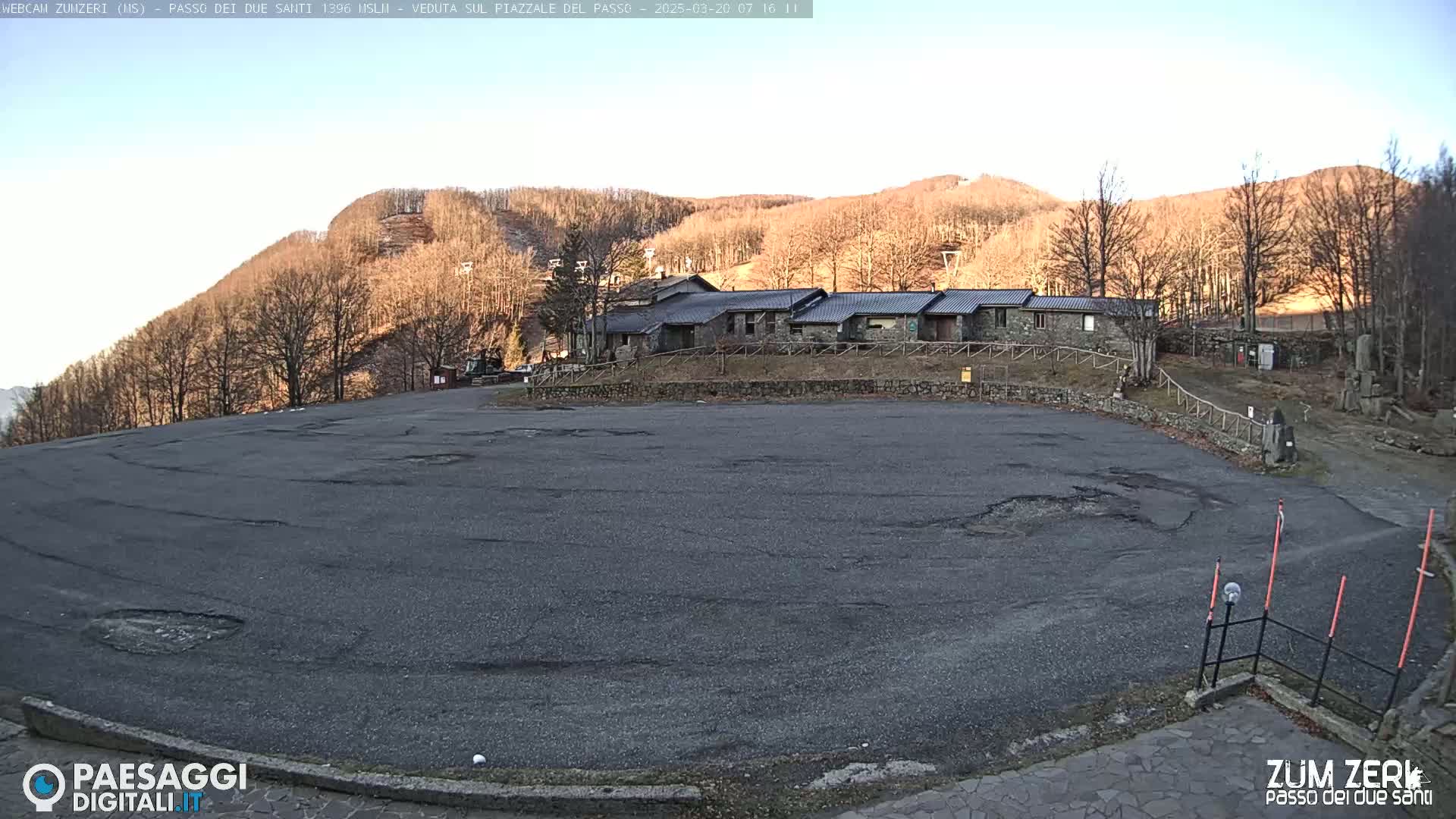 A large, empty asphalt parking lot with some potholes is situated in front of a stone building complex, all set against a backdrop of a hill covered in bare, brown trees under a clear, sunny sky.