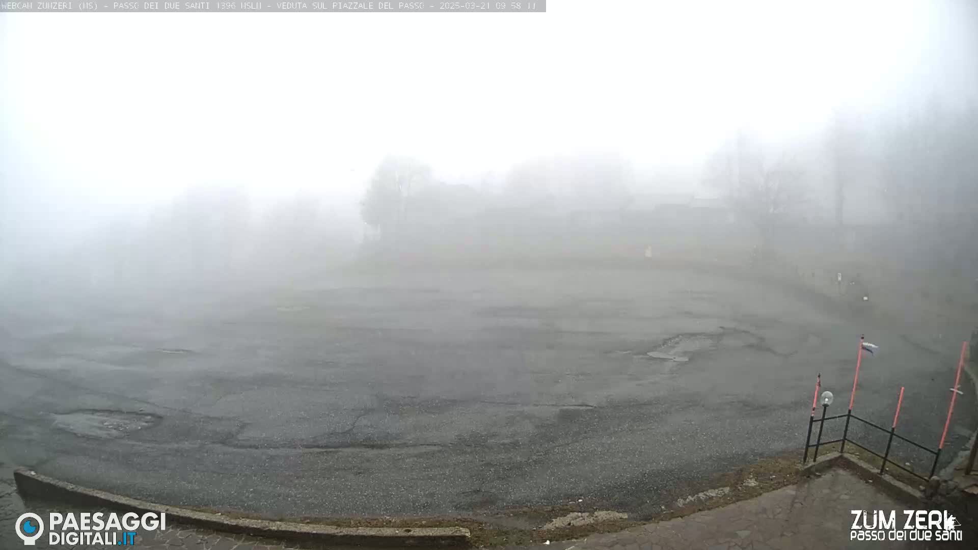 A foggy, overcast scene shows a large, empty asphalt area, partially bordered by a low curb and a metal fence, with some sparsely vegetated ground visible nearby.