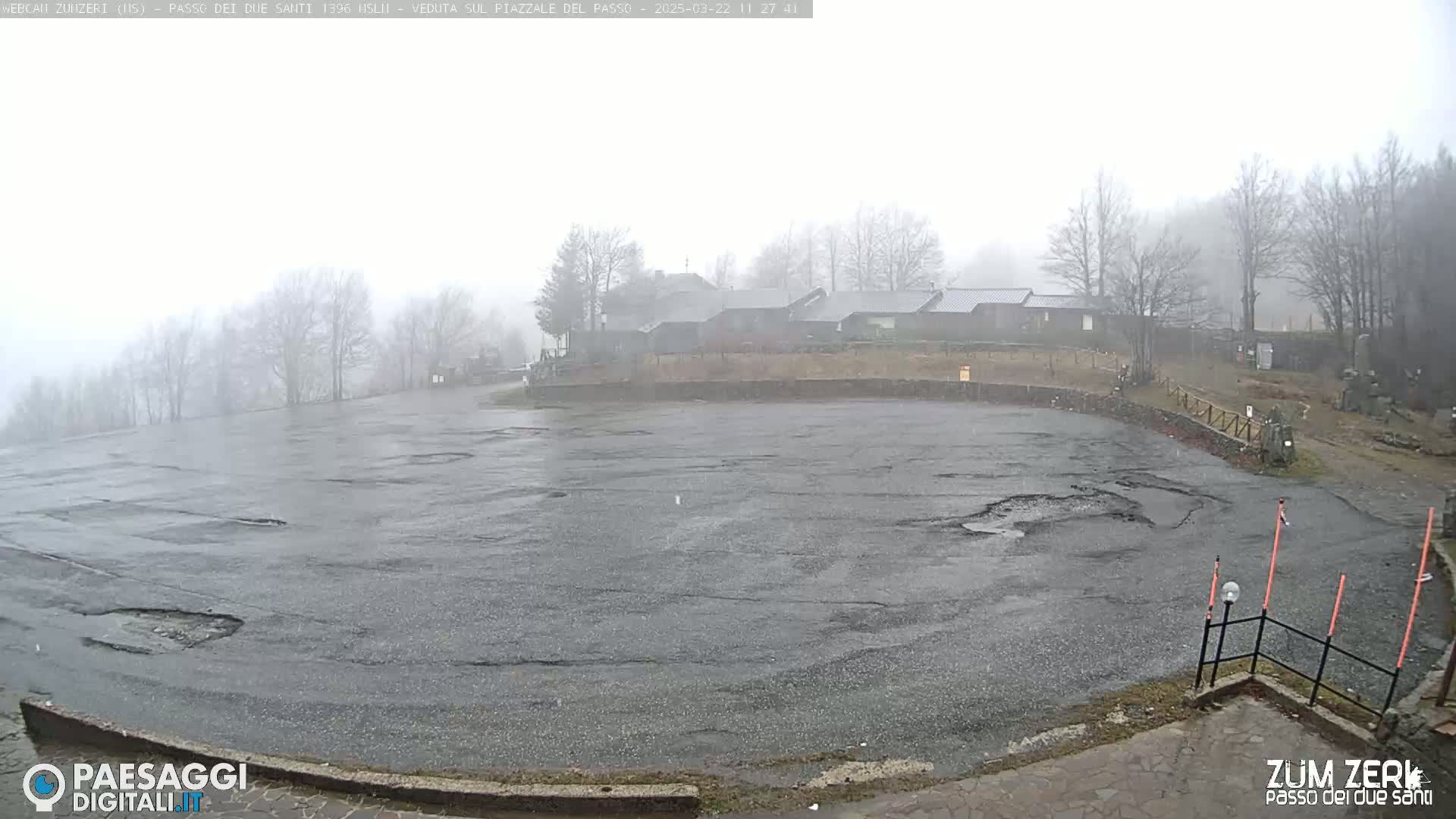 A large, rain-soaked asphalt parking lot is seen, with a line of buildings in the background under a foggy sky.