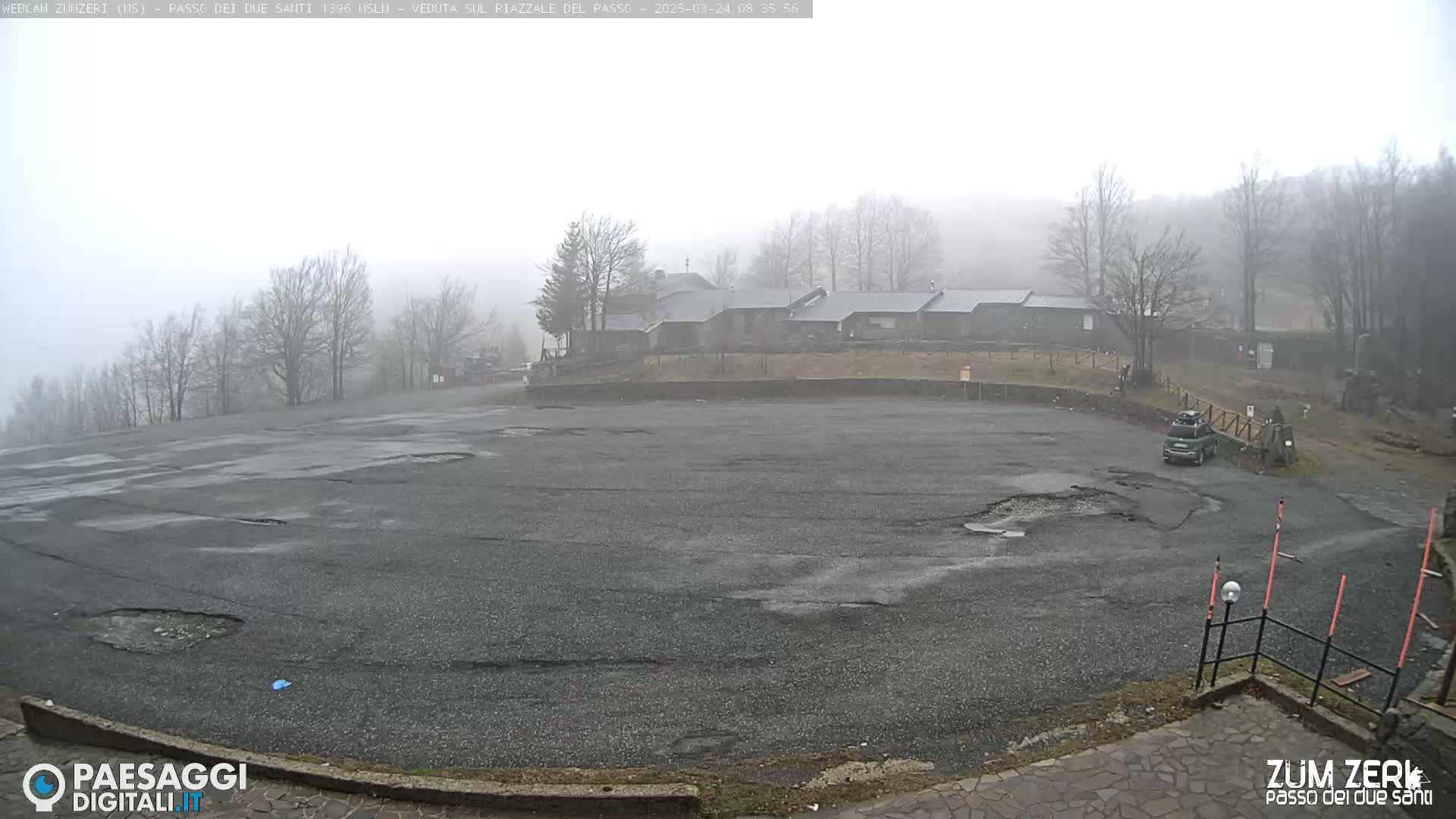 A foggy, overcast day reveals a mostly empty asphalt parking lot with potholes, a few bare trees, and a cluster of buildings in the background.