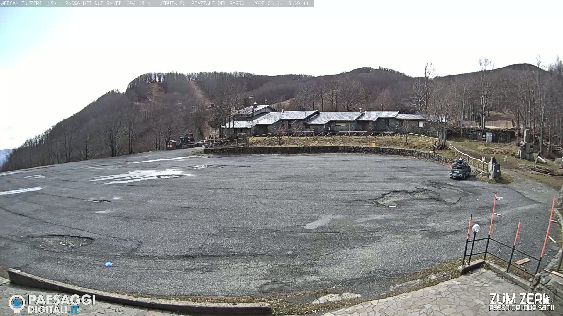 A large, empty asphalt parking lot sits in front of a stone building complex against a backdrop of a treeless, mountainous landscape under a mostly clear sky.